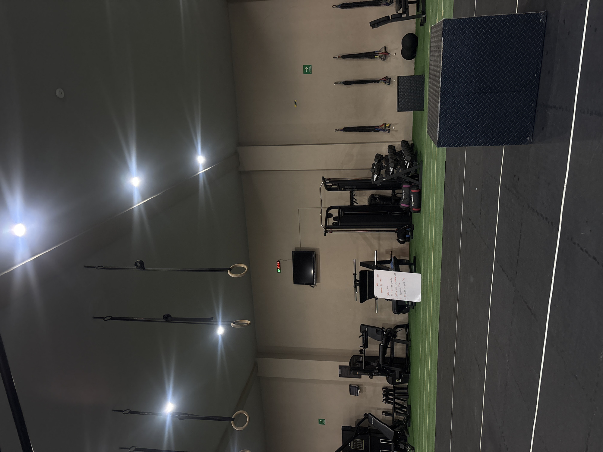 This media file presents an interior view of a well-equipped, empty gym or fitness center located in San Bernardino Tlaxcalancingo, Mexico. The space features light grey walls and a floor with distinct zones: a green artificial turf area and a black rubberized track marked with parallel white lines.
The gym is outfitted with various exercise equipment. On the right, there are racks filled with dumbbells, multiple strength training machines (including what appears to be leg press and leg extension/curl stations), and barbells. Two black plyometric boxes with a diamond plate texture are visible on the green turf, one larger than the other. Resistance bands are neatly hung on the wall. From the ceiling, several gymnastic rings are suspended, suggesting areas for bodyweight and functional training. Bright overhead artificial lights illuminate the room, casting noticeable glare. A smoke detector is also visible on the ceiling.
A small flat-screen television is mounted on the wall, and below it, a digital display shows "UP 27:51," likely indicating a timer or stopwatch. Nearby, a white sign or piece of paper outlines a workout, with partially readable text including "4," "300m run," "125 en sled push's," "3000/s/1000," "12," and "10 ager του μπρ." Green arrow signs pointing upwards are present on two different walls. The absence of people suggests the gym is unoccupied.
