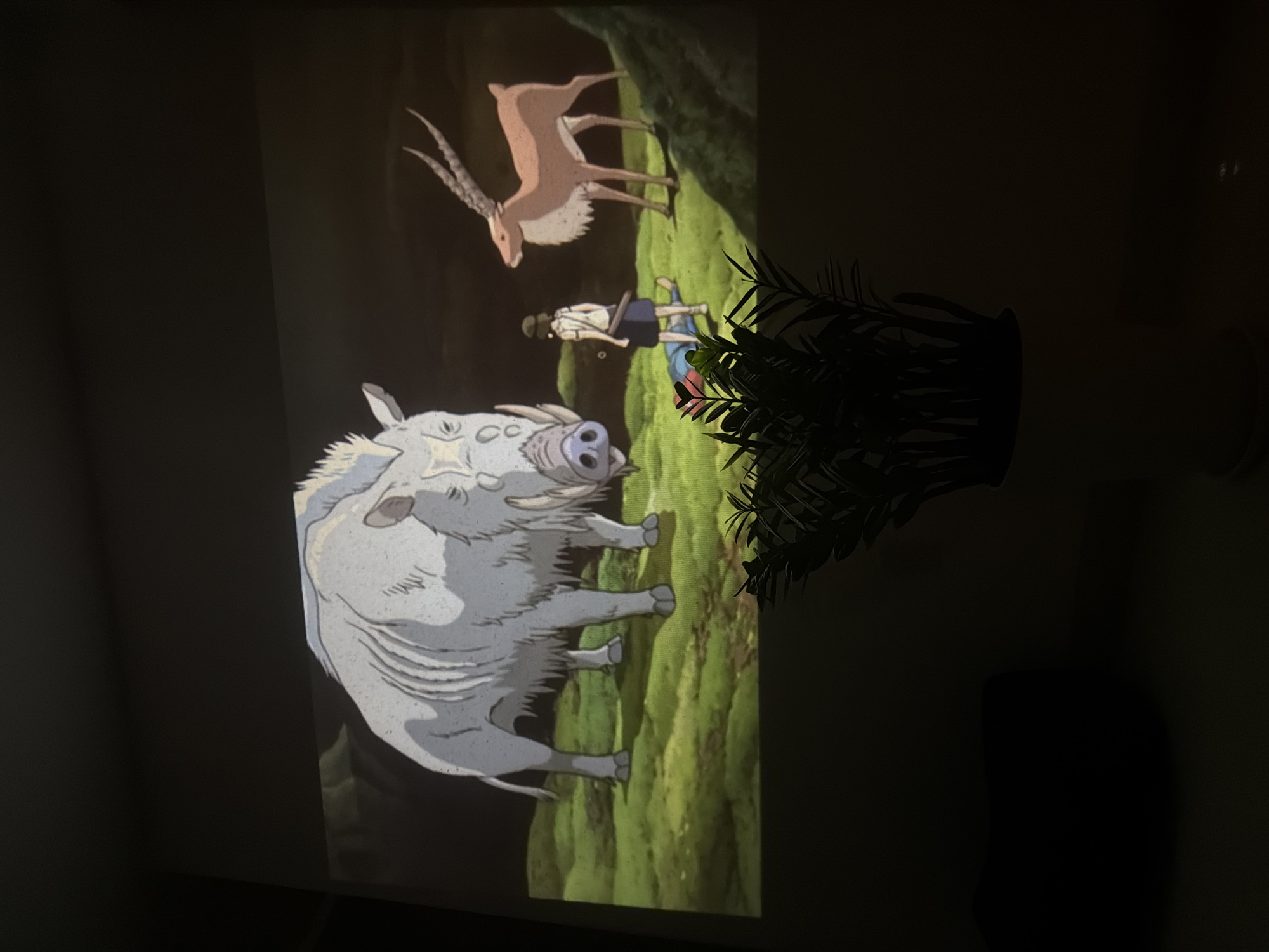 The image shows a projection of a scene from the animated film "Princess Mononoke". The scene features a large, white boar-like creature with tusks, a deer-like animal with antlers, and a human character lying on the ground. The background appears to be a forest or wooded area. A potted plant partially obscures the projection. The setting is unknown and is not necessarily located in the city of Đà Nẵng, Vietnam.