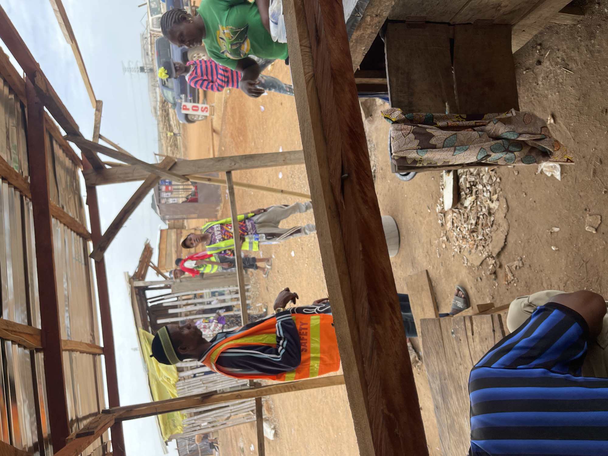 The image depicts an outdoor scene in what appears to be a construction or market area of Gosa, Nigeria. The foreground is dominated by rough, wooden structures, likely stalls or shelters, with raw lumber prominently featured. The ground is dirt with scattered debris. Several individuals are visible. One man wears a safety vest over striped clothing. Other individuals are seen wearing bright colored clothing. In the background, there are parked cars and other simple buildings. The overall impression is of a busy, somewhat disorganized informal commercial area.