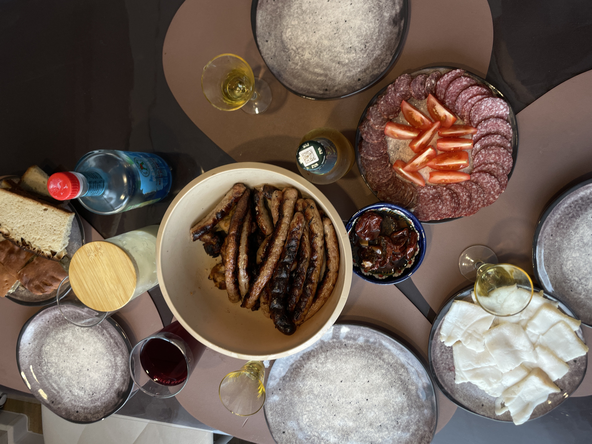 A top-down view shows a table laid out with food and drinks in what appears to be Подшивалово, Russia. There are five plates visible, each with a gray mottled pattern. One plate holds slices of bread. A large bowl in the center is filled with grilled sausages. Adjacent to the sausages is a small blue bowl containing what appears to be sundried tomatoes. Another plate presents sliced salami arranged around tomato wedges. A plate on the right has white, thinly sliced food, possibly cheese or fat. There are two bottles, one with a blue label and red cap, the other with a green cap and tan liquid. Several glasses are scattered across the table, filled with different colored liquids, including wine, and clear beverages. The table surface has a brown and gray pattern.