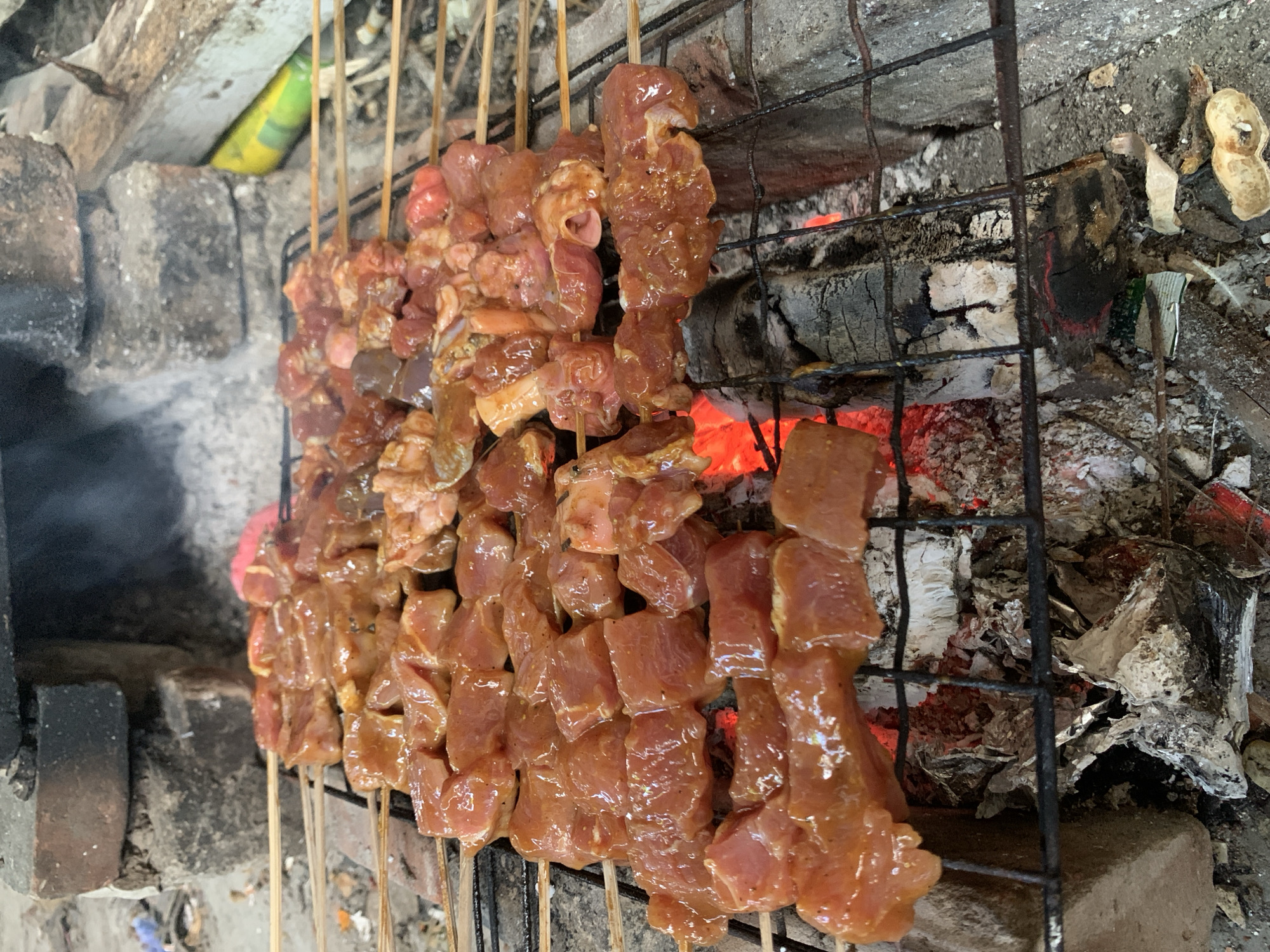 The image shows skewers of meat cooking on a charcoal grill. The meat is cut into cubes, coated in a marinade, and arranged on thin wooden skewers. The grill is positioned over hot coals, which are glowing red. Ash and debris are scattered around the edges of the grill. The grill appears to be improvised and made of iron bars. A building appears to be visible in the distance. This scene is likely in Karangkemiri, Indonesia.