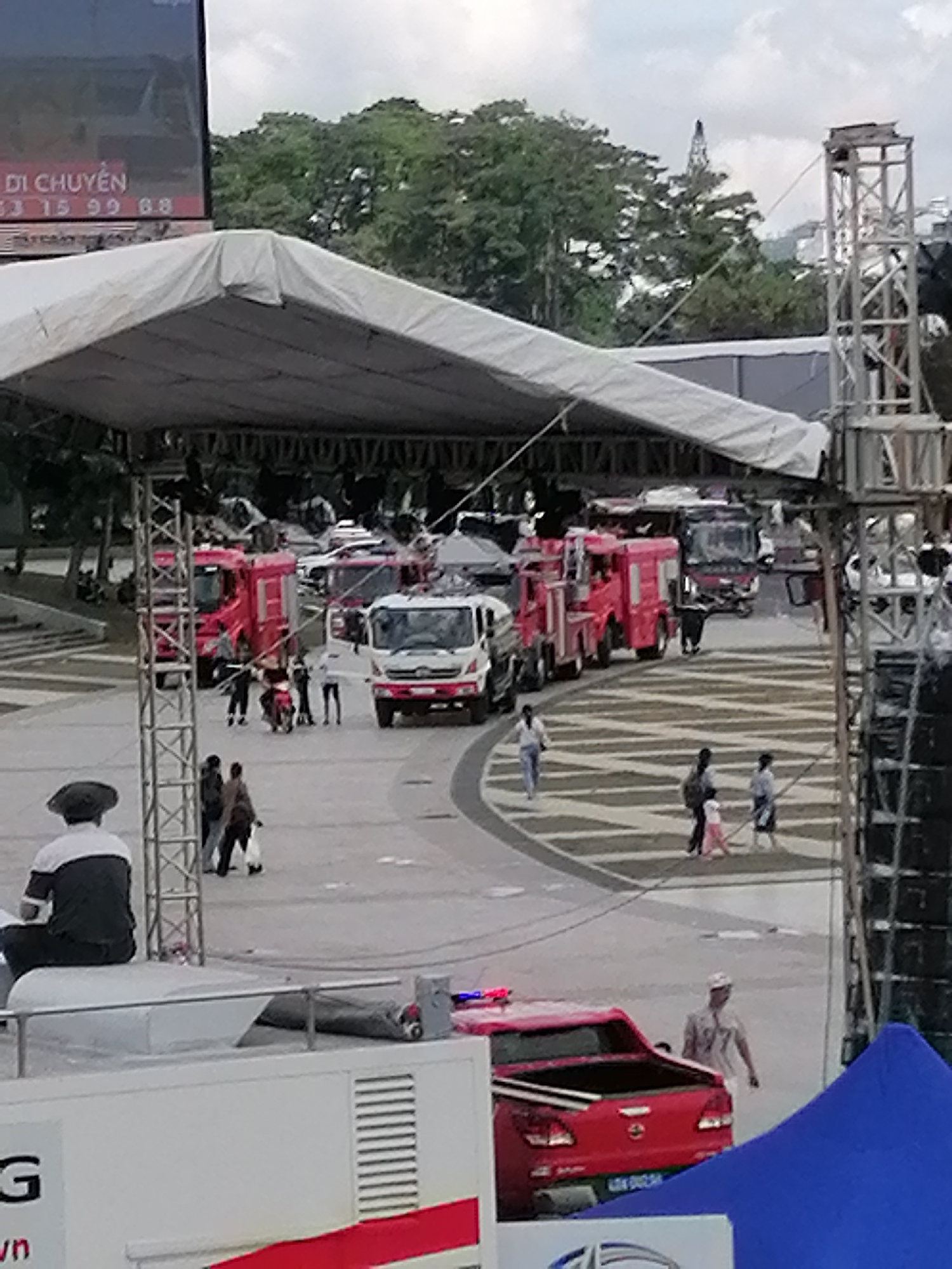 In Da Lat, Vietnam, an outdoor event is underway under a large white tent structure. Several fire trucks, one white and two red, are parked in the vicinity, suggesting a potential emergency response or a public exhibition. A red pickup truck with flashing lights is also present, further emphasizing a focus on emergency services. Numerous individuals are seen walking and interacting in the open space, some gathered in small groups. The scene is characterized by a mix of people, vehicles, and temporary structures, with a backdrop of lush green trees and a cloudy sky.