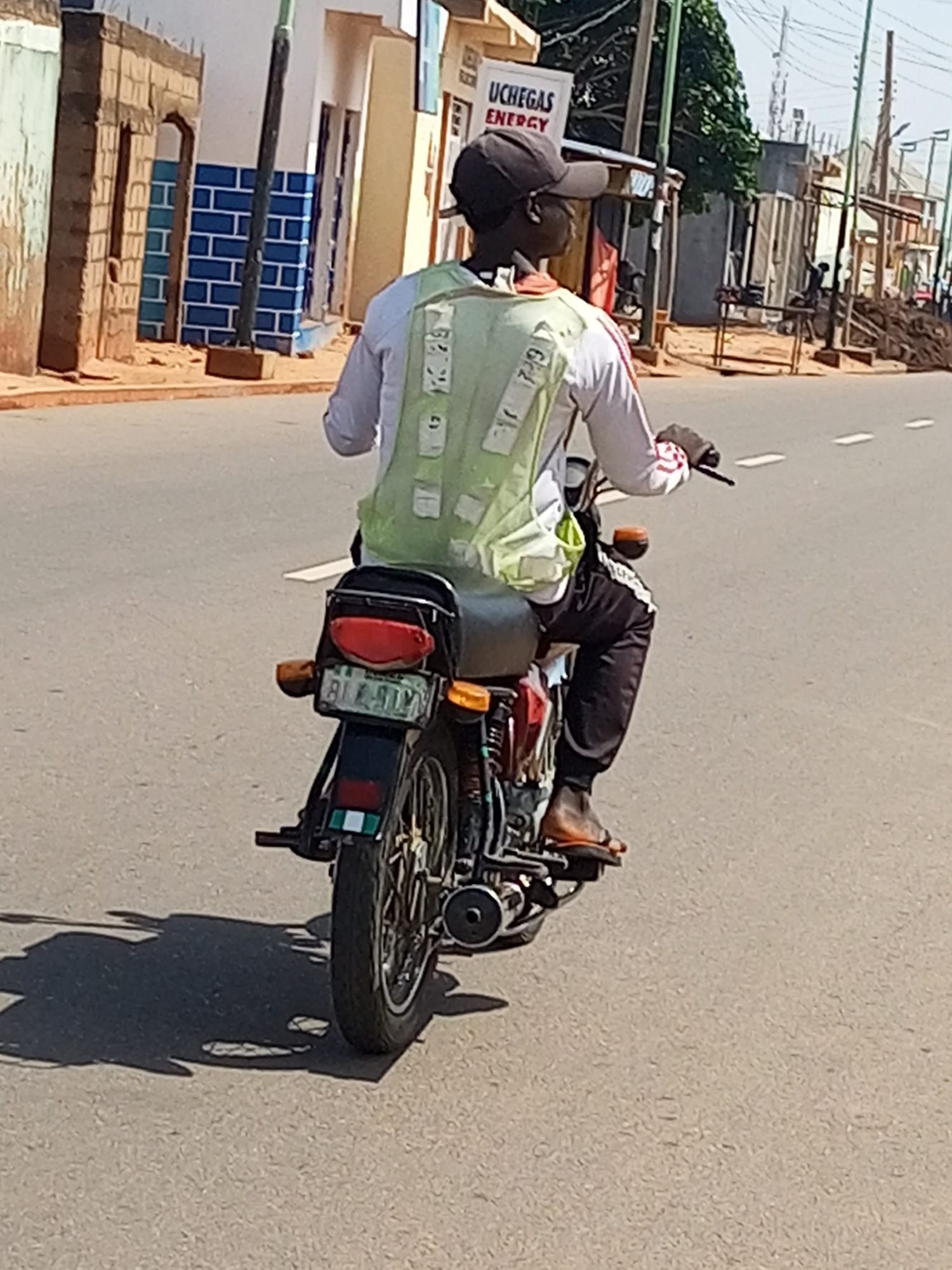 A person wearing a dark baseball cap and a light-colored long-sleeved shirt with red stripes on the sleeves is riding a motorcycle. They are wearing a bright green high-visibility vest over their shirt. The vest has white rectangular reflective panels. The rider is also wearing dark pants and brown sandals. The motorcycle is dark-colored with a red taillight. The license plate is visible and appears to have a Nigerian registration format. The motorcycle has a green and white Nigerian flag sticker on the rear. The rider's left foot is visible on the motorcycle's footrest. The motorcycle has a silver exhaust pipe and spokes. The scene takes place on a paved road with white dashed lane markings. In the background, there are buildings with brick and painted facades, some with blue tile accents. A sign for "UCHEGAS ENERGY" is visible on one of the buildings. Trees and utility poles are also present. The ground is asphalt. The overall lighting suggests a sunny day. The image appears to be taken in Gombe, Nigeria, based on the license plate and the style of buildings.