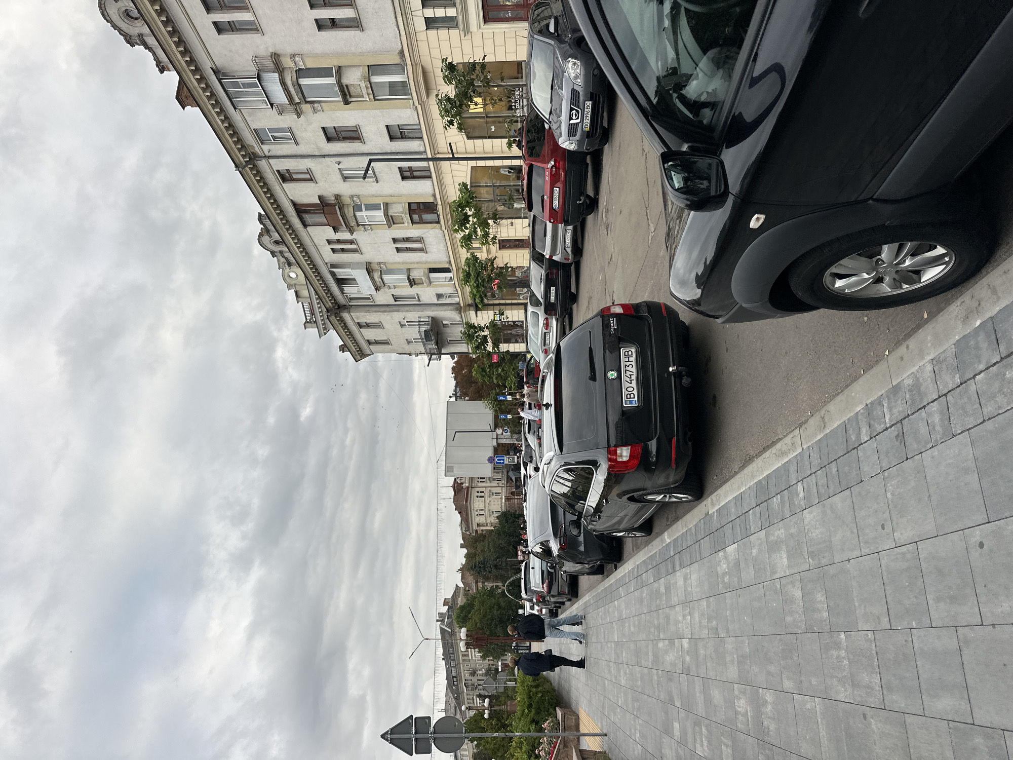 This image captures a street scene in Ternopil, Ukraine, under an overcast sky. A row of parked cars lines the right side of the street, with a pedestrian sidewalk on the left. The sidewalk is paved with grey rectangular tiles, bordered by a curb and a strip of smaller, darker grey tiles. Two individuals, a man and a woman, stand on the sidewalk, facing away from the camera and towards the parked cars. The man is wearing a dark jacket and blue jeans, while the woman is in a dark jacket and dark pants.
Further down the street, more cars are parked, including a black station wagon with license plate "BO 4473 HB" prominently visible. The buildings along the street are multi-story structures with traditional architecture, featuring ornate detailing along the rooflines and balconies. Some of the buildings have shops or businesses on the ground floor, indicated by storefront windows. Trees with green foliage are planted along the sidewalk and in front of some buildings. Traffic signs, including a triangular sign with a red border and a white background with black symbols, are visible on the left side of the street. The overall impression is a moderately busy urban environment.