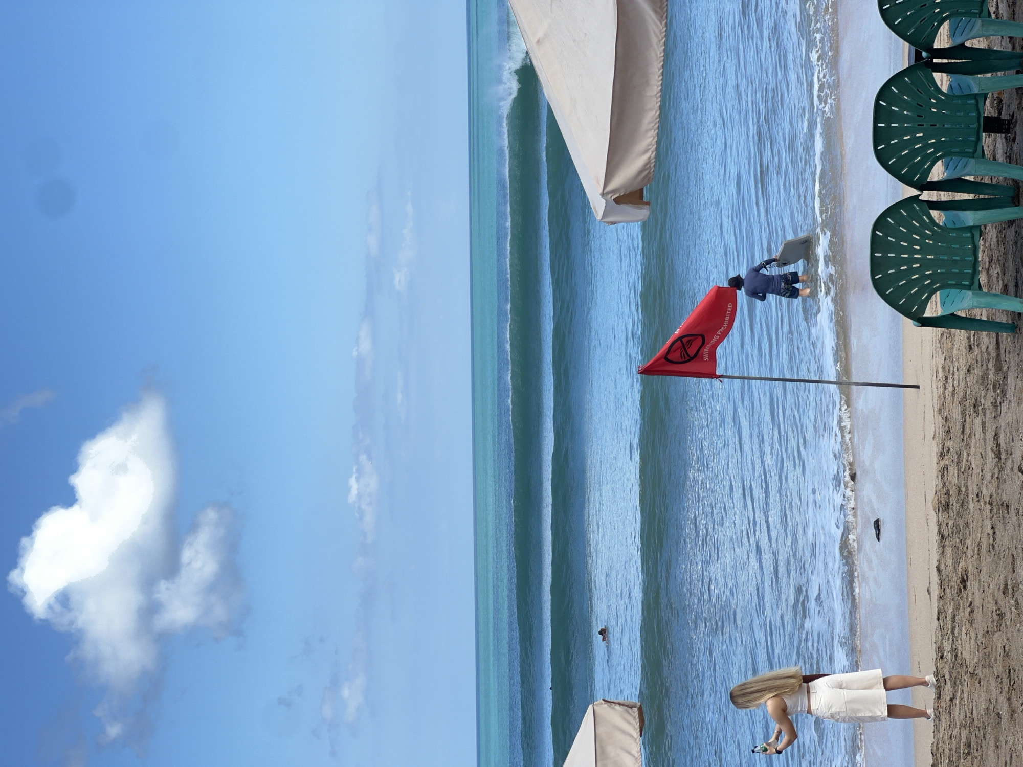 In Kuta, Indonesia, a wide shot of a beach shows the ocean with multiple sets of waves breaking. In the foreground, a woman with long blonde hair stands on the sand, holding a bottle and a phone as if taking a picture. She is wearing a white crop top and white shorts. To her right, a red flag with a prohibition symbol and text that reads "SWIMMING PROHIBITED" is planted in the sand, near the water's edge. A person in a dark blue shirt and patterned shorts is wading into the ocean, carrying a surfboard. Two large cream-colored beach umbrellas are visible, one on the left and one on the right, partially framing the scene. In the distance, several surfers are in the water, and a person is swimming. The sky is blue with scattered white clouds. In the immediate foreground, three green plastic chairs are placed on the sand.