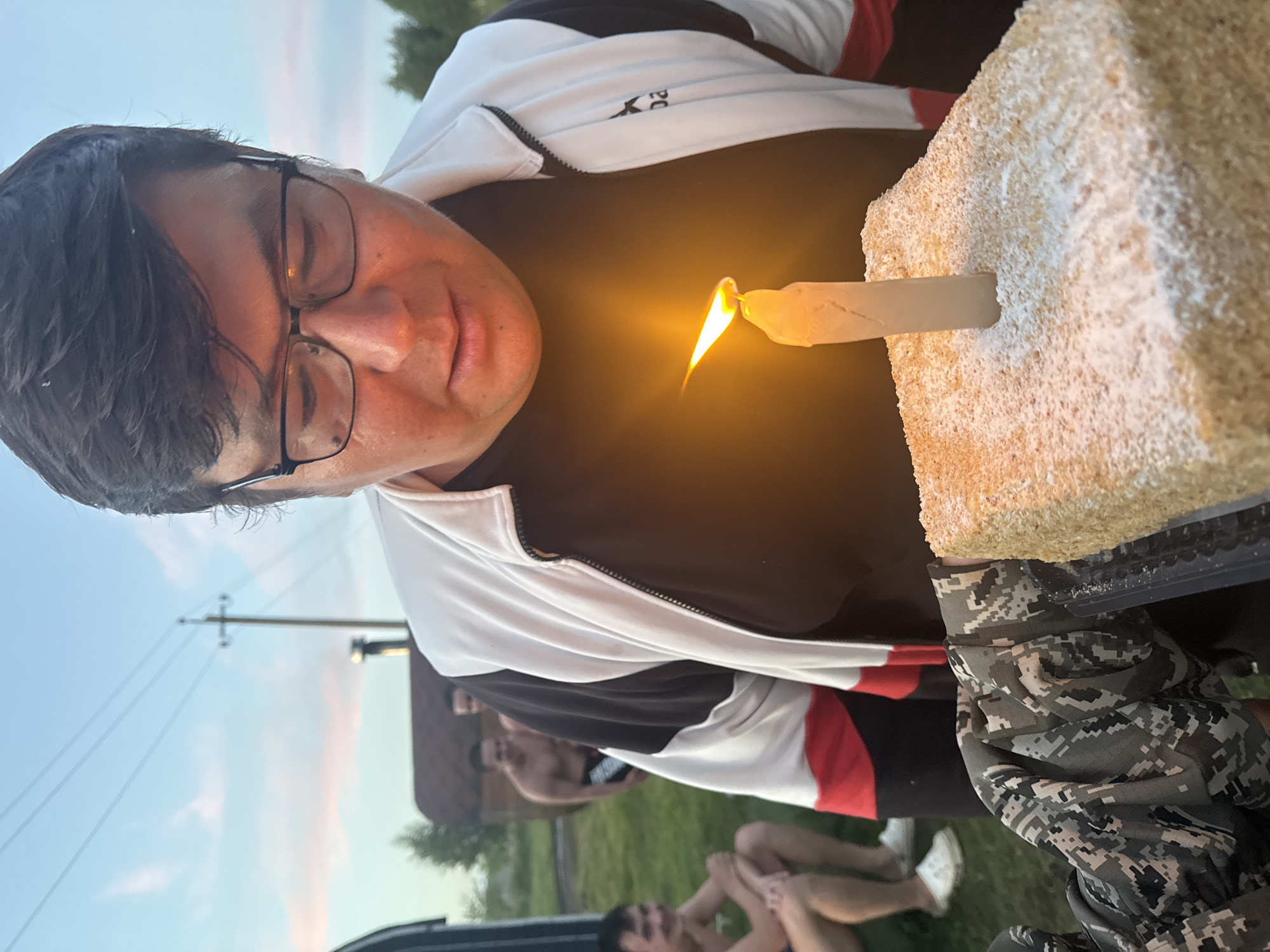 An outdoor, close-up shot shows a man in his 30s with dark hair and glasses blowing out a single lit candle on a rectangular cake. He is wearing a black t-shirt and a white and black jacket with red accents. The cake is dusted with powdered sugar. In the background, two other shirtless men are visible near a wooden structure, possibly a sauna, and some trees. The setting appears to be a rural or semi-rural area, consistent with the outskirts of Yakty-Kul, Russia, given the presence of natural elements and simple structures. The sky is overcast with a hint of sunset colors. The focus is on the man and the cake, with the background blurred.
