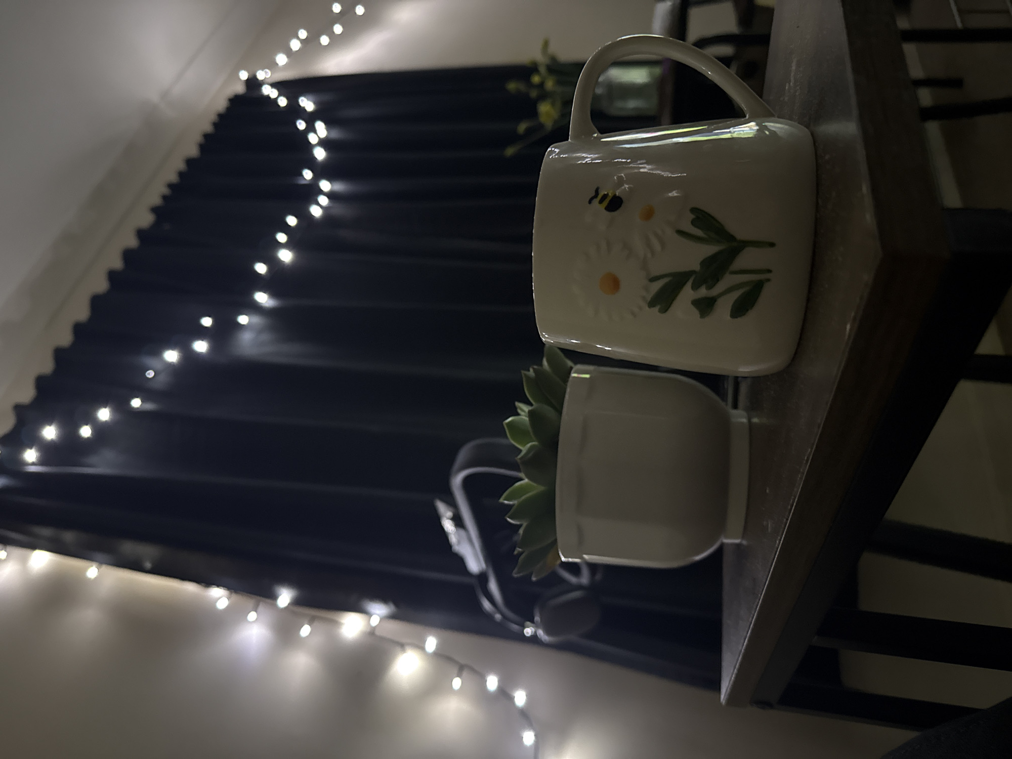 This indoor scene is located in Great Yarmouth, United Kingdom. The image depicts a dimly lit room, focusing on a dark wooden surface, likely a table or shelf, in the foreground. On this surface, two primary objects are present: a white ceramic pot containing a green succulent plant positioned to the left, and a white ceramic mug with a handle visible on the right, decorated with an embossed design of white daisies, yellow centers, green stems, and a small black and yellow bee.
In the midground, a pair of black over-ear headphones or a VR headset is partially visible, resting against or hanging near a set of dark, pleated curtains that occupy the central and right background. A string of small, warm white LED fairy lights is draped across the top edge of these curtains and extends along a light-colored wall on the left side of the frame. Further into the background, to the right of the mug, a blurry glass vase containing light-colored flowers, possibly yellow, is faintly discernible on another surface. The overall lighting is low, originating primarily from the fairy lights, casting a soft glow on the immediate surroundings. No individuals are present in the image, and no active interactions or movements are depicted.