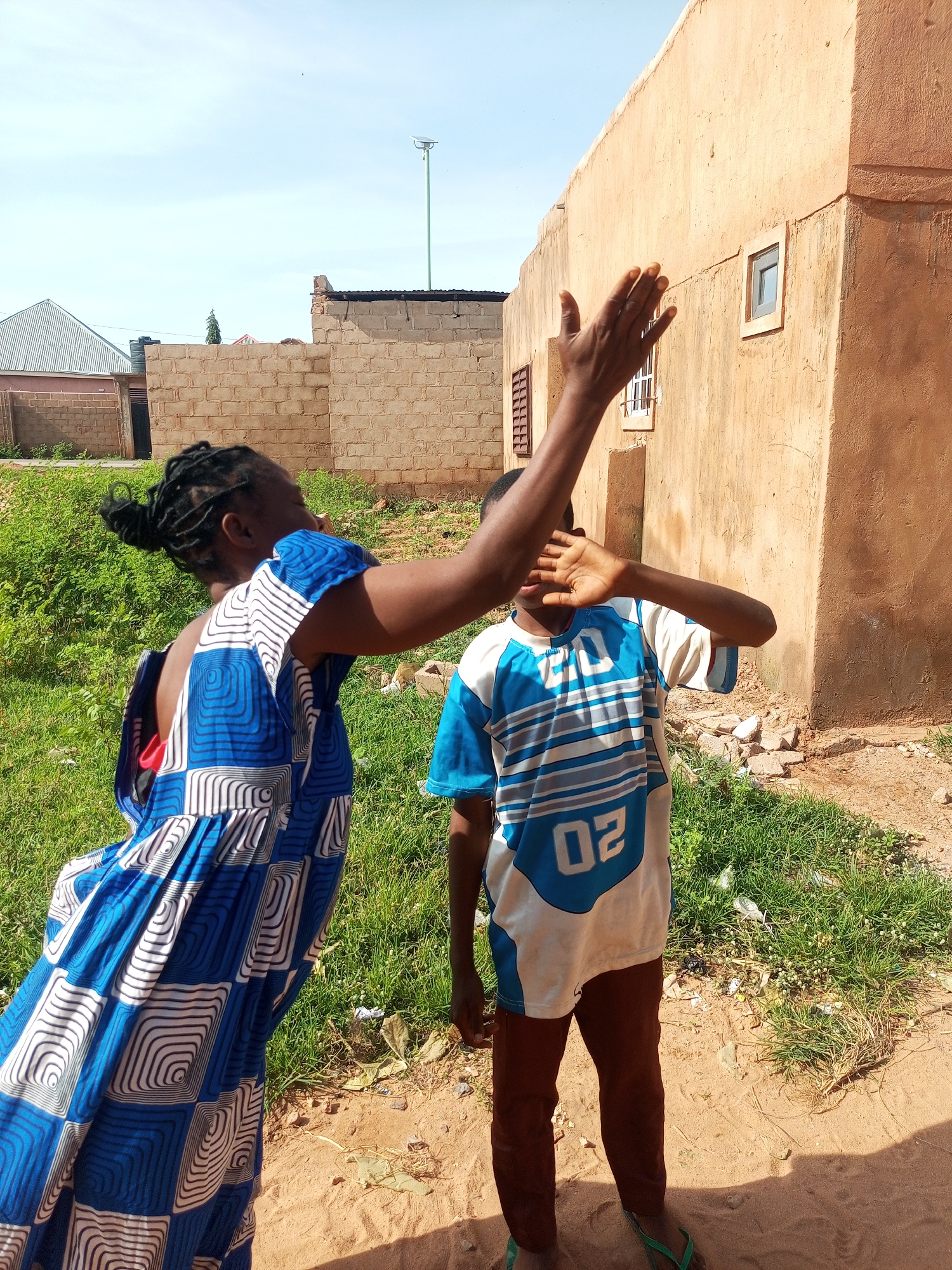In Tempure, Nigeria, two individuals are captured in an outdoor scene near a rustic, tan-colored building. The building, constructed with mud bricks, features a few small, square windows and a weathered facade. In the foreground, a person wearing a vibrant blue and white patterned dress, with their hair in braids, extends an arm with an open hand towards the camera, as if to block it. Positioned slightly behind and to the right, a second individual, dressed in a blue and white striped t-shirt with the number "02" on it and dark brown trousers, shields their face with both hands. The ground is a mix of sandy earth and patches of green grass, with scattered debris. In the background, a lower brick wall with a tin roof is visible, along with a glimpse of another building with a corrugated metal roof. The sky above is a clear, light blue.