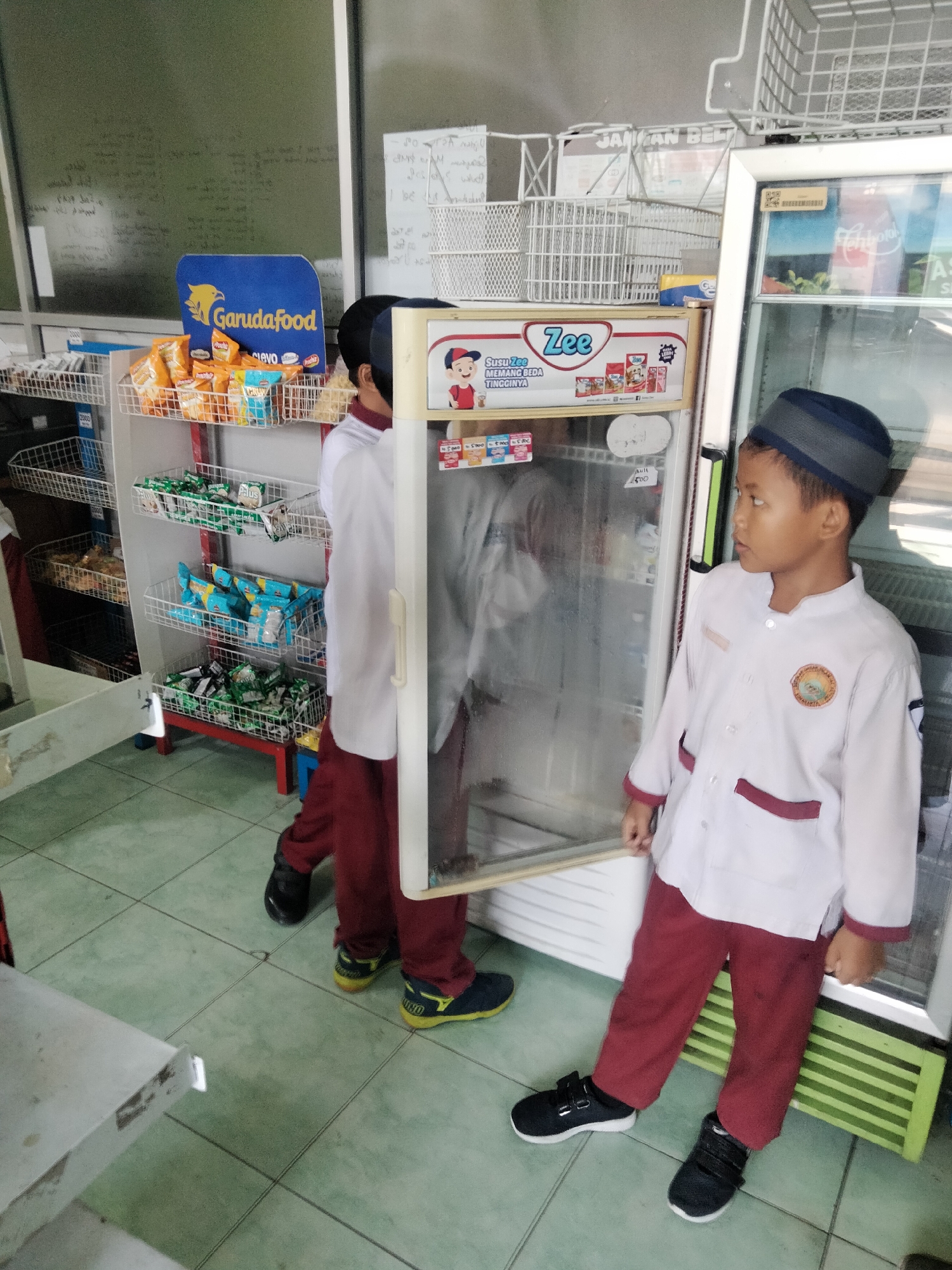The image depicts two young boys in school uniforms standing in front of a convenience store or school canteen. The boy on the right, closer to the camera, is wearing a white shirt with maroon trim and pants, and a dark blue and grey peci. He is looking to his right. The boy on the left, partially obscured by a glass-fronted refrigerator, is wearing a similar uniform.
The background shows shelves stocked with various snacks and drinks. A prominent blue sign with a yellow bird and the text "Garudafood" is visible on one of the shelves, indicating a brand of snacks. A large, white refrigerator unit with glass doors is in the center of the image, displaying products inside. The floor is tiled in a light green pattern. The overall setting suggests a retail or institutional environment, likely in Indonesia given the clothing and branding. The presence of school uniforms and the type of merchandise strongly suggests a school canteen or a small shop catering to students in Surakarta, Indonesia.