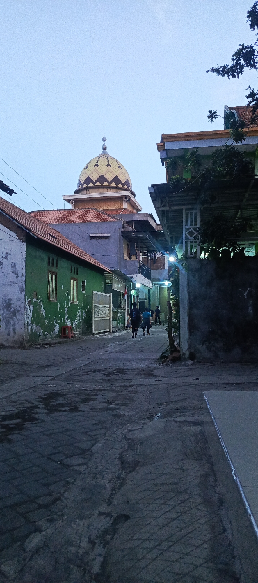 The image depicts a street scene at dusk in Tempel, Indonesia. The sky is a pale blue. A prominent golden-domed mosque is visible in the background, with a tiled roof on a surrounding building. In the foreground, a narrow street paved with interlocking blocks is lined with buildings.
On the left, a weathered green building with peeling paint has windows and an orange stool nearby. A large, ornamental gate is also present.
On the right, a building with a dark exterior wall is partly obscured by trees. A light-colored table or surface is visible in the lower right corner.
Several individuals are walking away from the camera down the street. The lighting suggests it is either early morning or late evening. The environment appears to be a residential or mixed-use area.