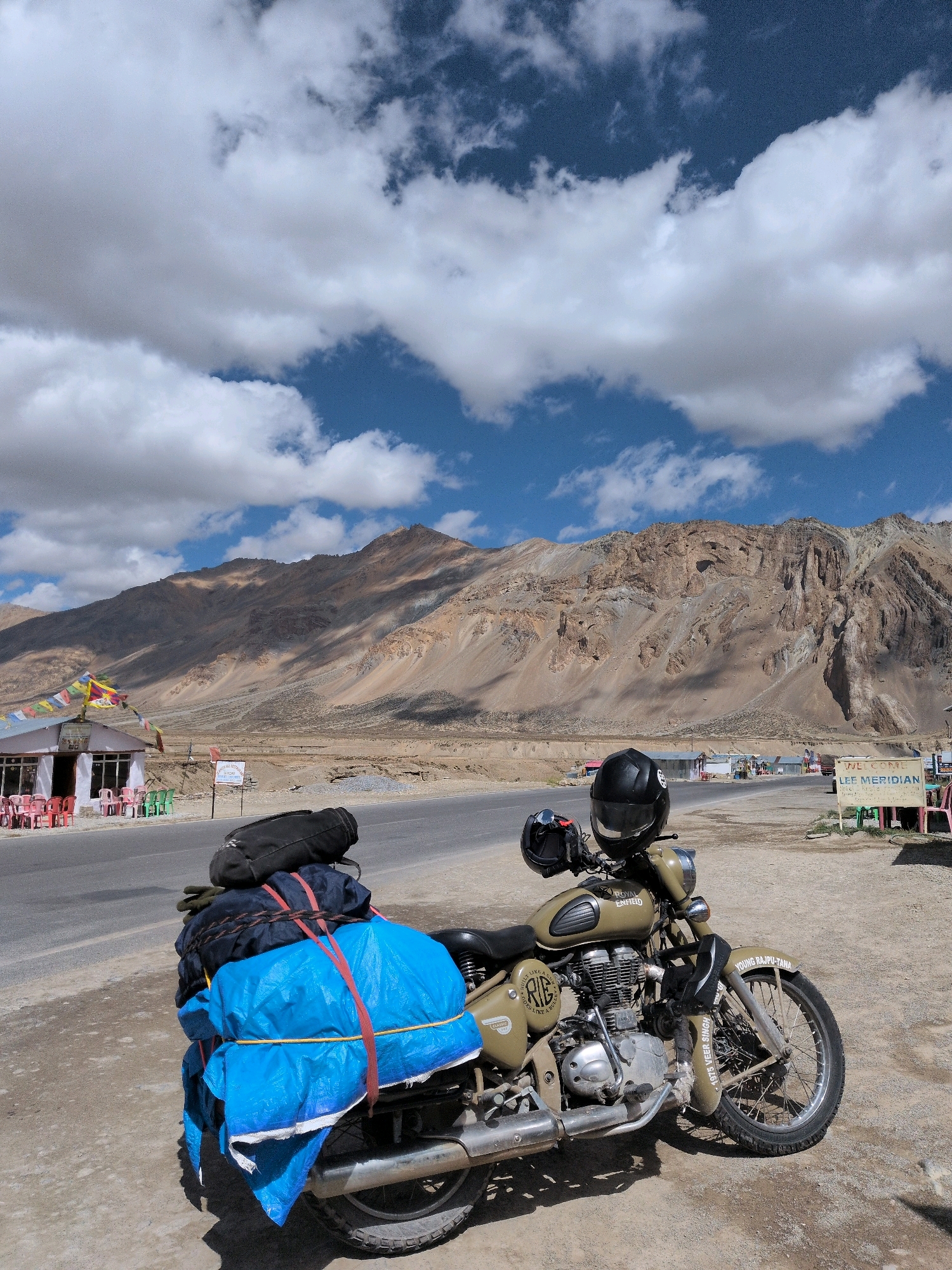 The image depicts a parked motorcycle loaded with luggage, situated on the side of a road in a mountainous, arid region. The motorcycle is a Royal Enfield, olive green in color, with a black helmet secured to its handlebars. A large blue tarp covers a significant portion of the luggage strapped to the rear of the motorcycle. In the background, a range of barren, rocky mountains dominates the landscape under a partly cloudy sky. To the left, a small building with colorful chairs outside, adorned with prayer flags, suggests a roadside establishment. A sign that reads "LEE MERIDIAN" is visible in the mid-ground. The overall scene conveys a sense of travel and remoteness.
