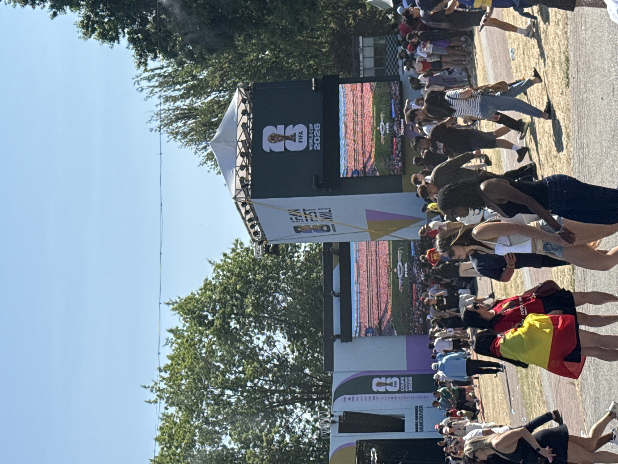 The image depicts a daytime outdoor scene at a FIFA Fan Festival event in Vancouver, Canada. A large, multi-sectioned structure with screens and branding is the central focus. The left section displays "FIFA FAN FEST 2026" and "COPE DU MONDE 2026" along with "VANCOUVER". The right section features a large screen showing a stadium filled with spectators, with a "FIFA WORLD CUP 2026" logo prominently displayed above it. A crowd of diverse individuals of varying ages and ethnicities are gathered in front of and around these structures. Many are casually dressed, with some wearing sports-themed clothing or carrying national flags, such as one person draped in a Spanish flag. People are walking, standing, and interacting, suggesting a lively public event. The environment appears to be a grassy or dirt area, with trees visible in the background and a clear blue sky overhead.