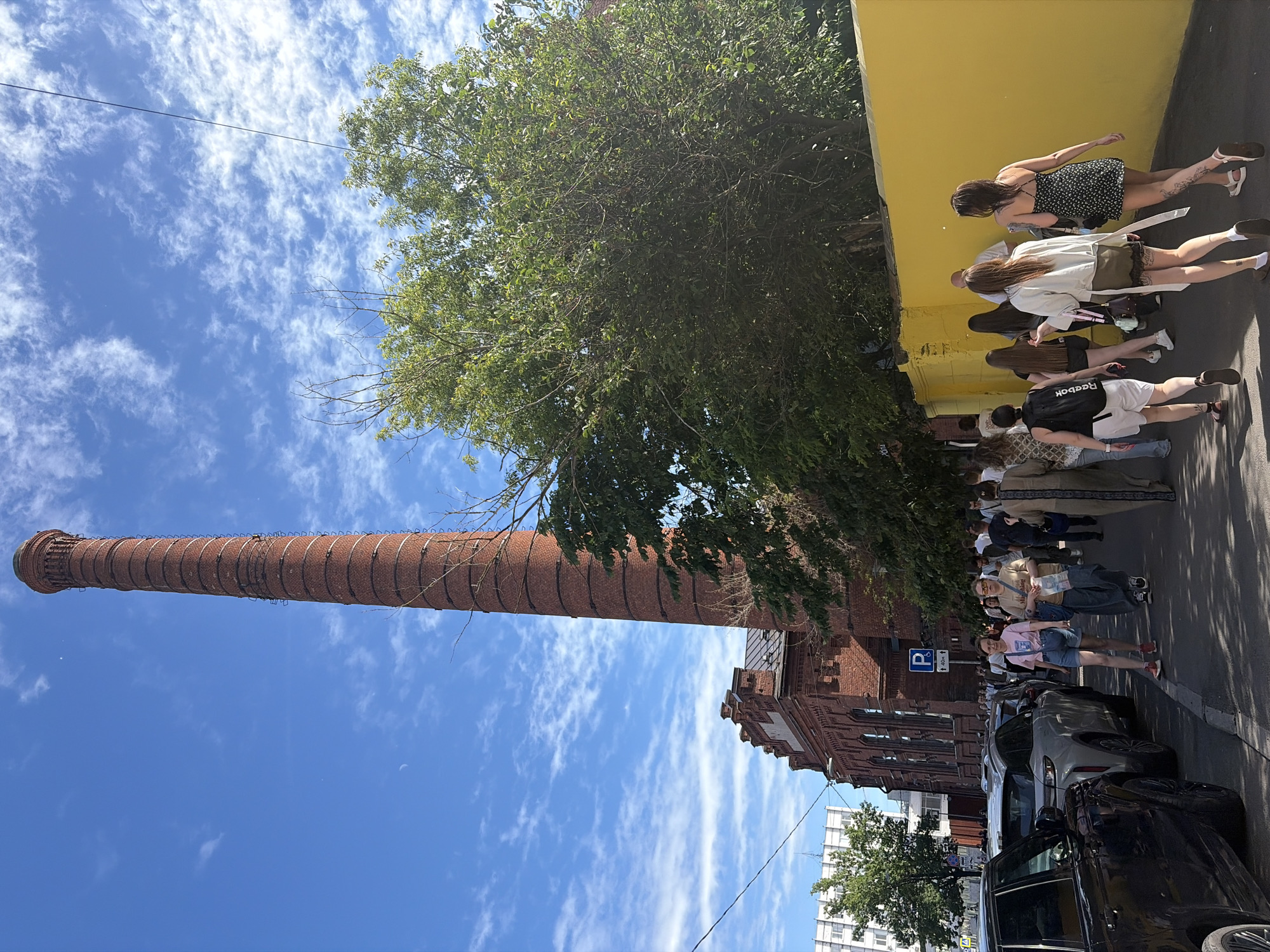 In Saint Petersburg, Russia, a group of people walk along a sidewalk beside a tall, brick industrial smokestack and a red brick building with an arched window facade. A bright yellow wall runs along the right side of the sidewalk. Several parked cars are visible on the left side of the image, including a black SUV, a silver SUV, and a white car. A parking sign with a "P" symbol and an arrow pointing upwards is mounted on a pole near the red brick building. The sky is blue with scattered white clouds. The trees in the image are full of green leaves. The people are dressed in casual summer clothing. Some are walking in the same direction, suggesting they are part of a group or event. The overall scene appears to be a sunny day in an urban area with historical industrial architecture.