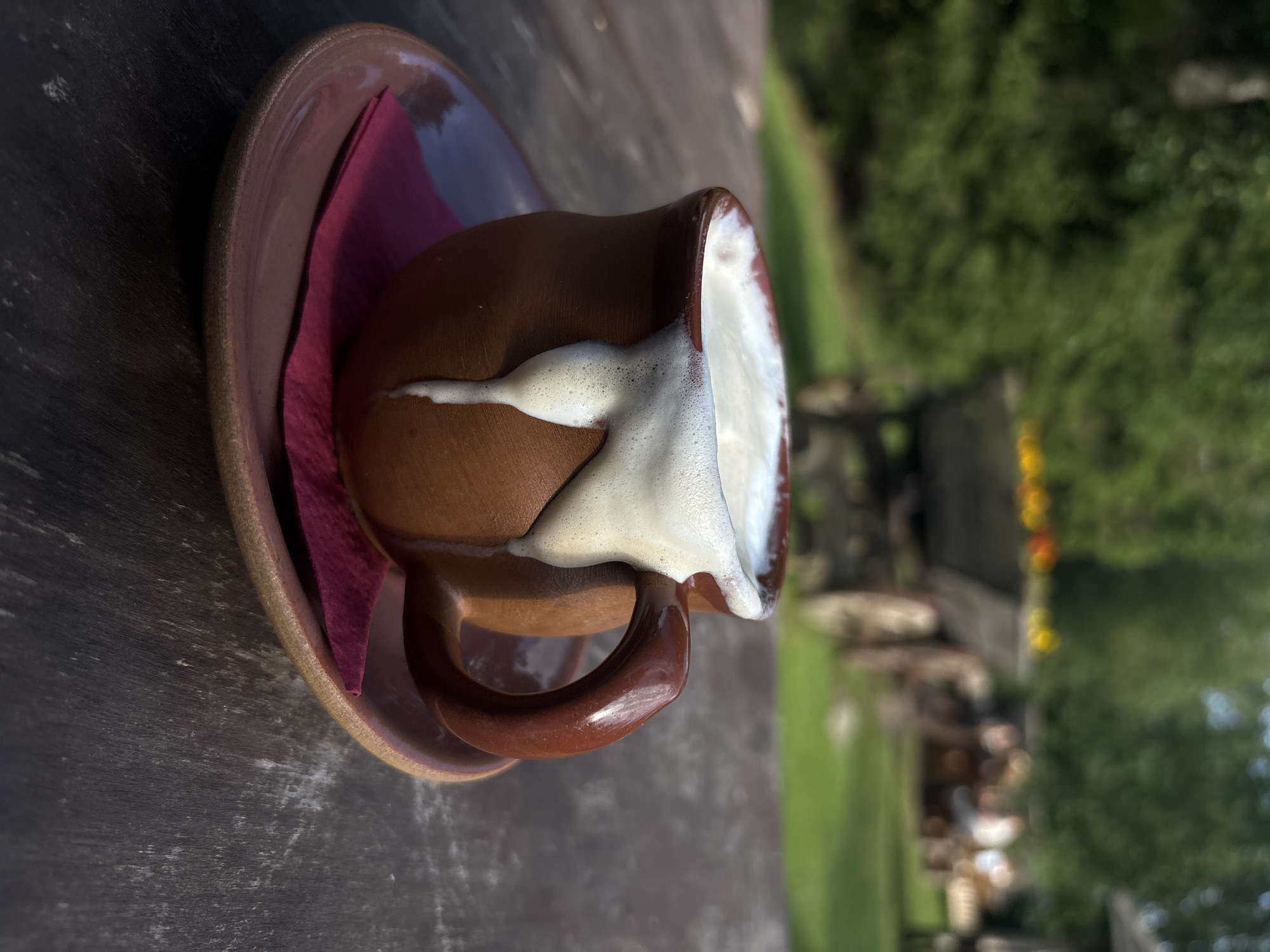 A close-up, eye-level shot focuses on a rustic, earthenware mug filled with a frothy, white beverage, likely coffee or milk, sitting on a dark wooden table. The mug has a thick, curved handle and is placed on a matching saucer, beneath which is a folded, dark pink napkin. The beverage has overflowed the rim of the mug, with a thick, foamy stream dripping down its side, creating a visually appealing texture.
The background is softly blurred, suggesting an outdoor setting in Saula, Estonia. Green foliage from trees forms a backdrop, with hints of sunlight filtering through. In the mid-ground, a weathered wooden cart, possibly an old horse-drawn cart, is visible, laden with what appear to be colorful flowers, adding a touch of rustic charm. Further in the background, blurred figures of people are seated at tables, hinting at a relaxed, outdoor dining or gathering area. The overall ambiance is serene and natural, with a focus on the simple pleasure of the beverage.