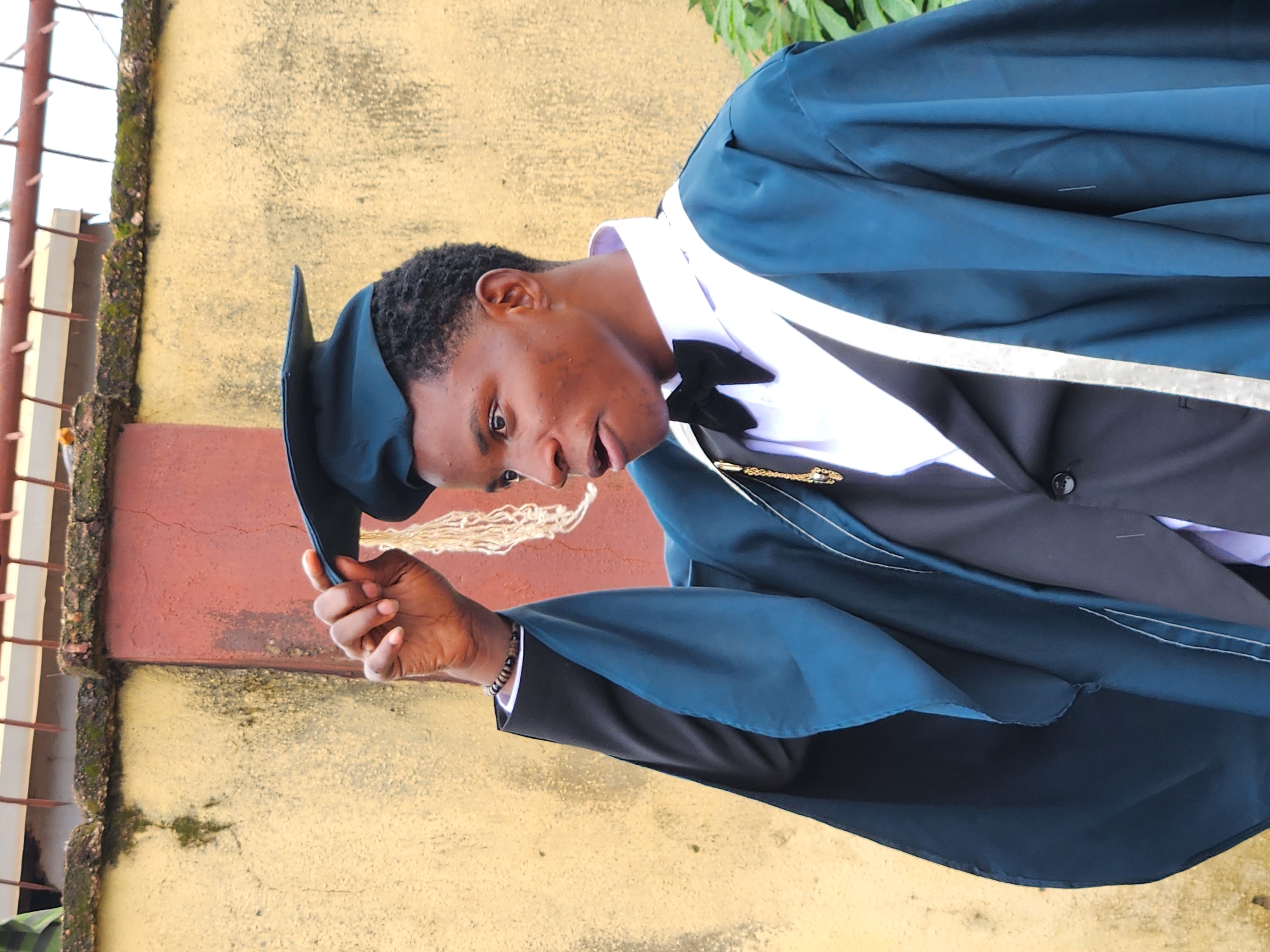 A young Black male graduate, wearing a dark teal graduation gown over a black suit with a white shirt and black bowtie, stands outdoors, holding the tassel of his graduation cap. The background is a textured yellow and red wall with a rusty metal railing at the top. The graduate is looking to his right with his mouth slightly open. The scene appears to be in Sabon Tasha, Nigeria, possibly during a graduation ceremony.