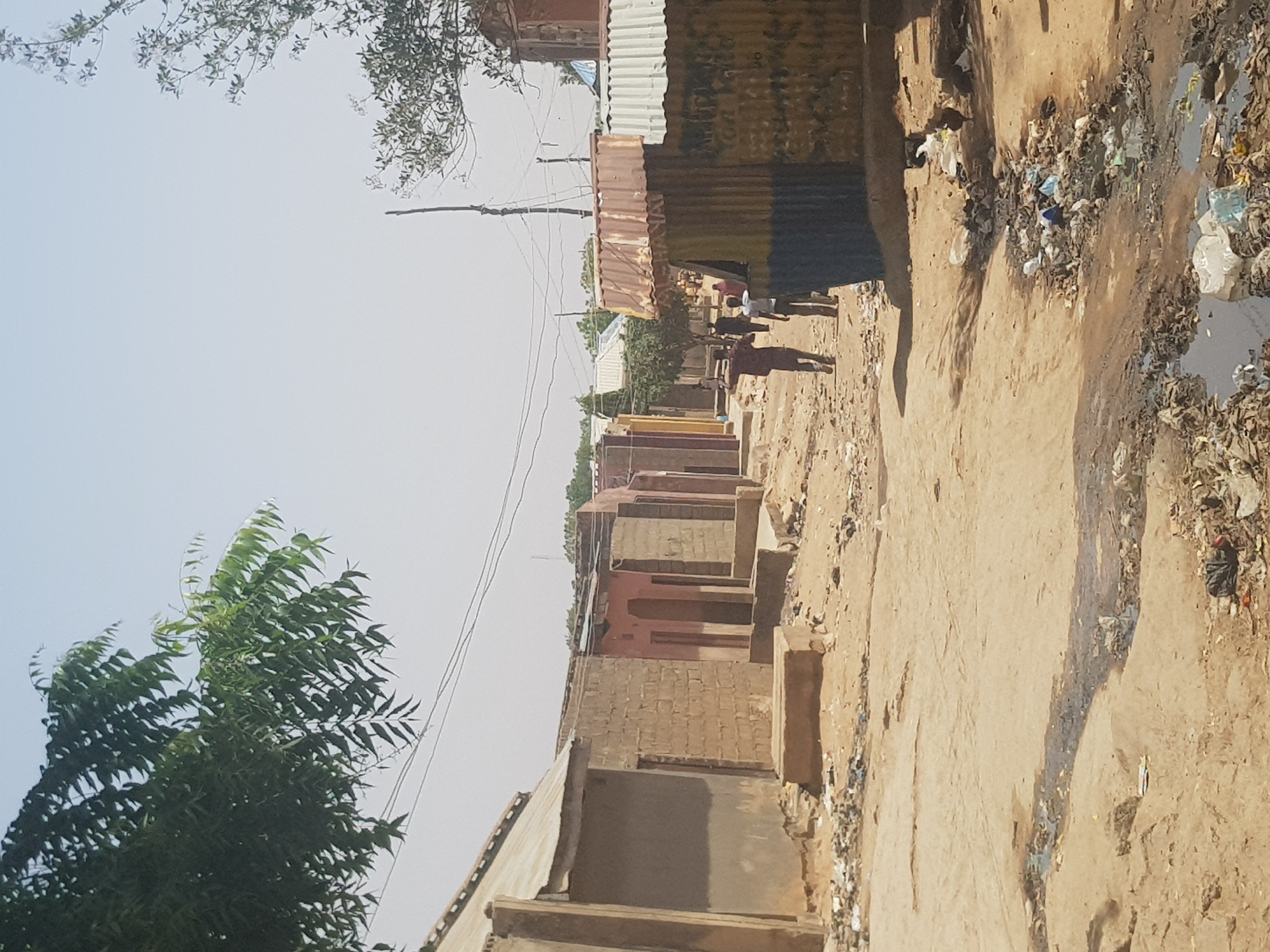 The image depicts a street in Dutsinma B, Nigeria. It shows a dirt road bordered by dwellings of varying construction, some appearing to be made of brick, others of corrugated metal. Litter and debris are visible along the roadside and in the drainage ditch. A group of people is walking down the street. Overhead power lines are strung above. Foliage from trees is visible in the foreground.