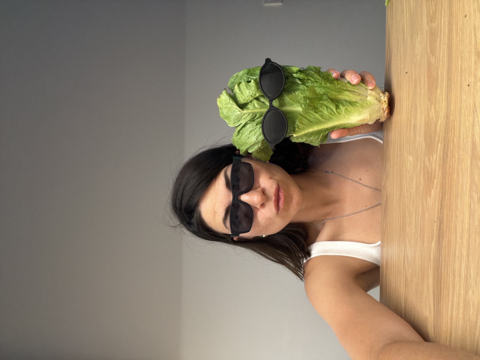 A woman is shown from a low angle. She wears a white tank top and a silver chain necklace. She has dark hair and sunglasses. She is holding a head of romaine lettuce that has sunglasses on top of it. The background is a plain gray wall on the left, and wood paneling on the right. The location cannot be determined from the image.