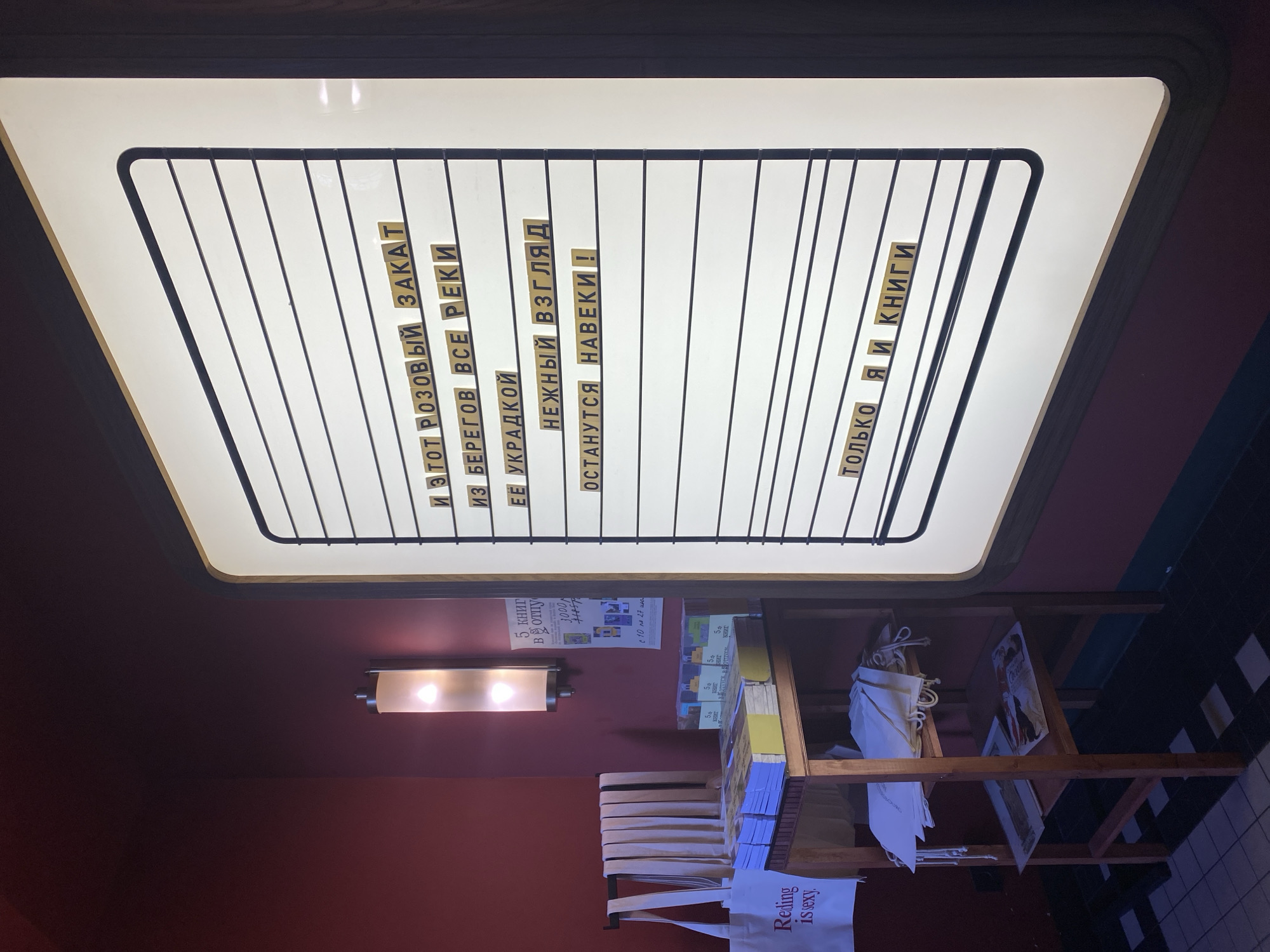 The image depicts the interior of a bookstore or cultural space, possibly located in Saint Petersburg, Russia, given the Cyrillic text visible. The dominant feature is a large, illuminated message board with a white background and black horizontal lines, displaying text composed of individual yellow tiles with black Cyrillic letters. The message on the board reads: "И ЭТОТ РОЗОВЫЙ ЗАКАТ ИЗ БЕРЕГОВ ВСЕ РЕКИ ЕЕ УКРАДКОЙ НЕЖНЫЙ ВЗГЛЯД ОСТАНУТСЯ НАВЕКИ ! ТОЛЬКО Я И КНИГИ". To the left of the message board, a wooden shelving unit holds stacks of books, some of which are wrapped in blue and yellow covers. Several white tote bags hang on a rack to the left of the shelving unit, with one bag prominently displaying the text "Reading is sexy." The walls are painted a deep red, and a vertically oriented light fixture is mounted on the wall. The floor is tiled with a pattern of dark and lighter squares. The overall atmosphere suggests a cozy and intellectually stimulating environment.