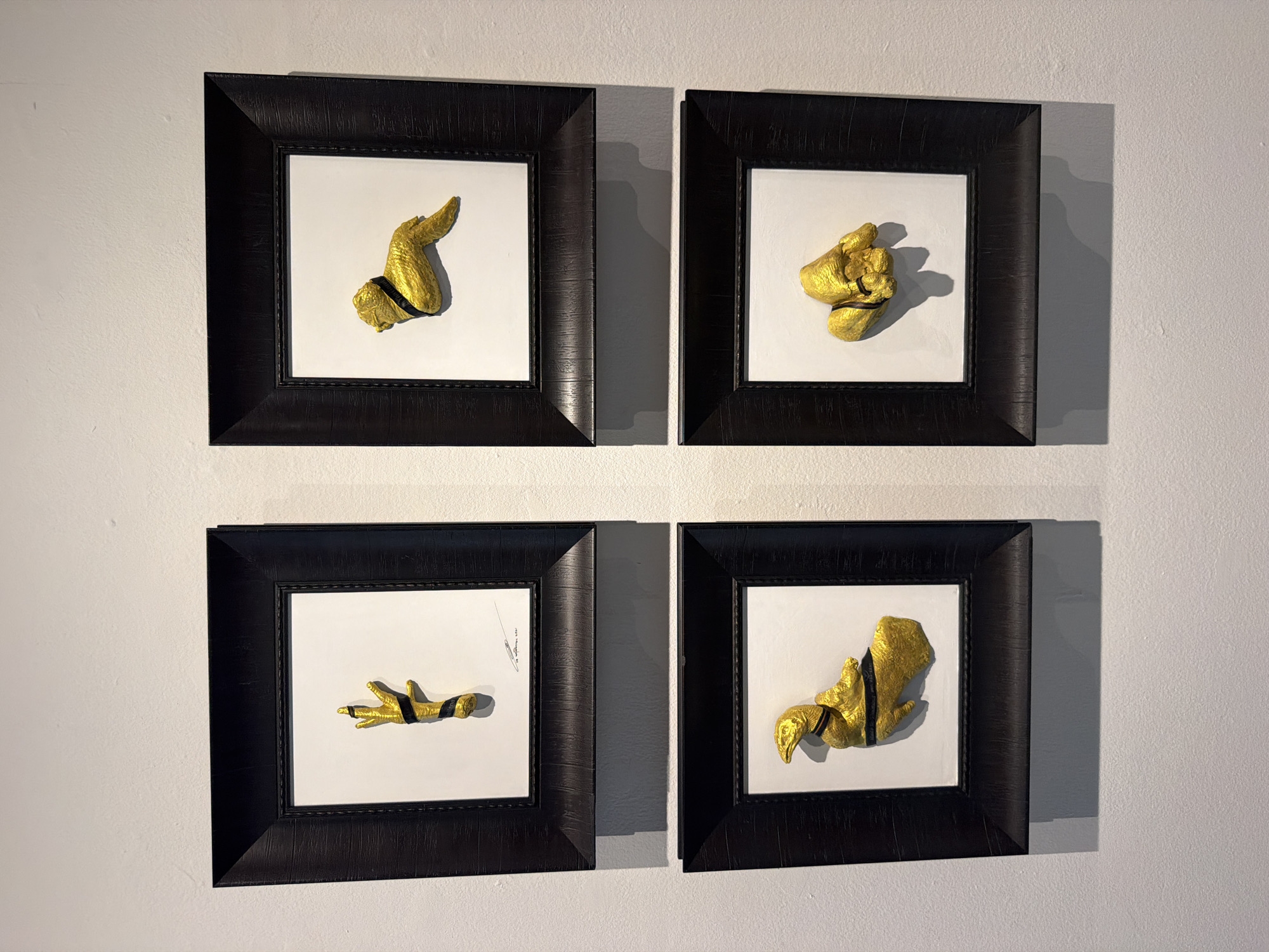 The image displays four framed artworks arranged in a two-by-two grid on a white wall. Each frame is black and has a textured surface. Inside each frame, a golden, three-dimensional object is mounted on a white background. The objects appear to be stylized representations of body parts.
The top-left artwork features a golden hand with fingers extended, wrapped in a black band. Below the hand, there is a signature and a date, "16 september, 2021".
The top-right artwork displays a golden object resembling a winged creature or a stylized limb, also with a black band.
The bottom-left artwork shows a golden object that appears to be a bird's head and neck, with a body that is partially visible and wrapped in a black band.
The bottom-right artwork contains a golden object that looks like a clenched fist or a folded hand, with a black band around it.
The overall presentation suggests an art exhibition or gallery setting. The artwork is displayed in a well-lit environment, with shadows cast on the wall below the frames, indicating a light source from above. The wall is a plain white, textured surface, typical of gallery walls. There are no people visible in the image. The specific location in Bangkok, Thailand, cannot be determined from the visual information provided in the image.