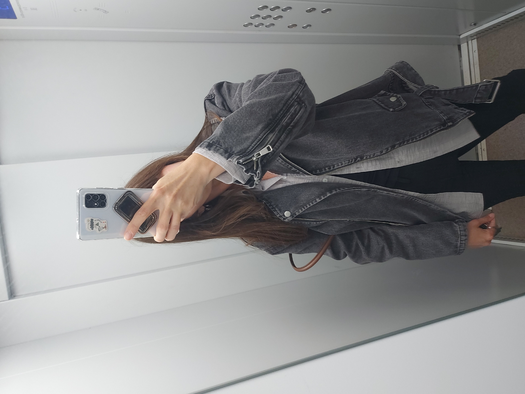 A mirror selfie captures a person with long brown hair, whose face is obscured by a light-colored smartphone held up for the photo. The individual wears a dark gray denim-style jacket with a zippered sleeve and a belt, over a light-colored collared shirt and dark pants. A brown strap, possibly from a bag, is visible over one shoulder. The smartphone features multiple rear cameras, a transparent case adorned with small cartoon-like stickers (one depicting a stylized figure), and a black phone grip. A ring is visible on one of the person's fingers.
The setting appears to be an indoor space, likely an elevator or a narrow hallway, characterized by light-colored, reflective walls and ceiling. The ceiling shows several clusters of small, circular metallic elements, possibly indicator lights or buttons. The reflection of the floor is also light-colored and reflective. The activity is taking a casual self-portrait. No specific time of day or weather is discernible, and the person's expression is not visible. The location context is Chaikovsky, Russia.
