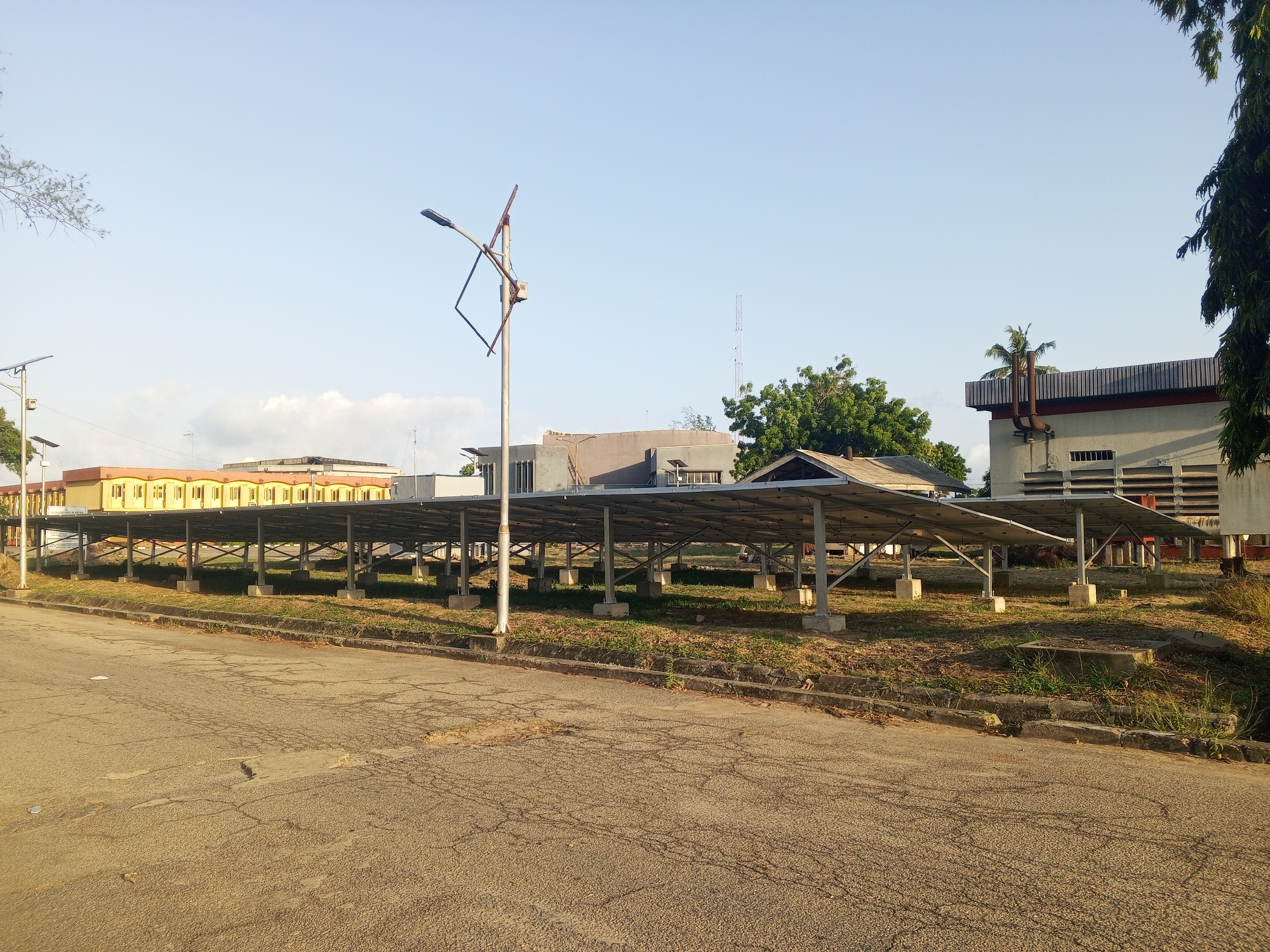 The image depicts a street scene in Tawpo, Nigeria, featuring a row of solar panels erected on concrete supports. The panels are situated on a grassy area adjacent to the road. Behind the panels are several buildings of varying architectural styles, including one with a distinct yellow facade. A street lamp stands near the panels, its design appearing incomplete. The road surface is cracked and shows signs of wear. The sky is clear and light blue, suggesting a daytime scene. Vegetation, including trees and grasses, are visible throughout the environment.