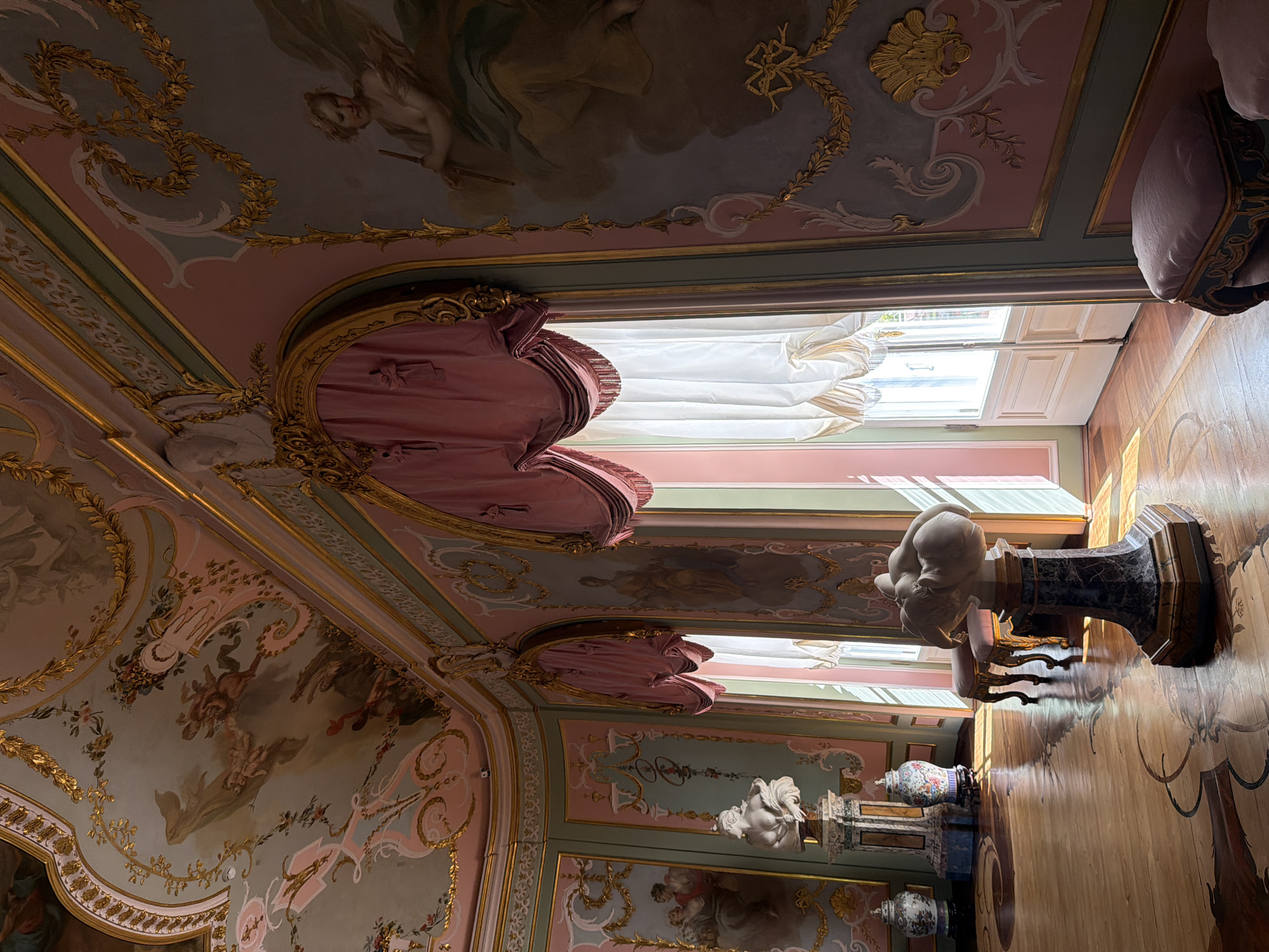 This is a medium-wide interior shot of a grand room with ornately decorated walls and ceiling, a polished wooden floor with inlaid patterns, and sunlight streaming in from two large windows. The walls are painted in pastel colors of pink and light green, with elaborate gold trim and moldings. Several large murals adorn the walls, depicting mythological figures and cherubic beings. Above, the ceiling is also heavily decorated with frescoes and gilded accents. Two large windows with draped pink valances and sheer white curtains are visible, letting in natural light. On the floor, there are two marble busts on pedestals, a pair of antique vases, and two small, upholstered stools. The overall impression is one of opulence and historical grandeur.