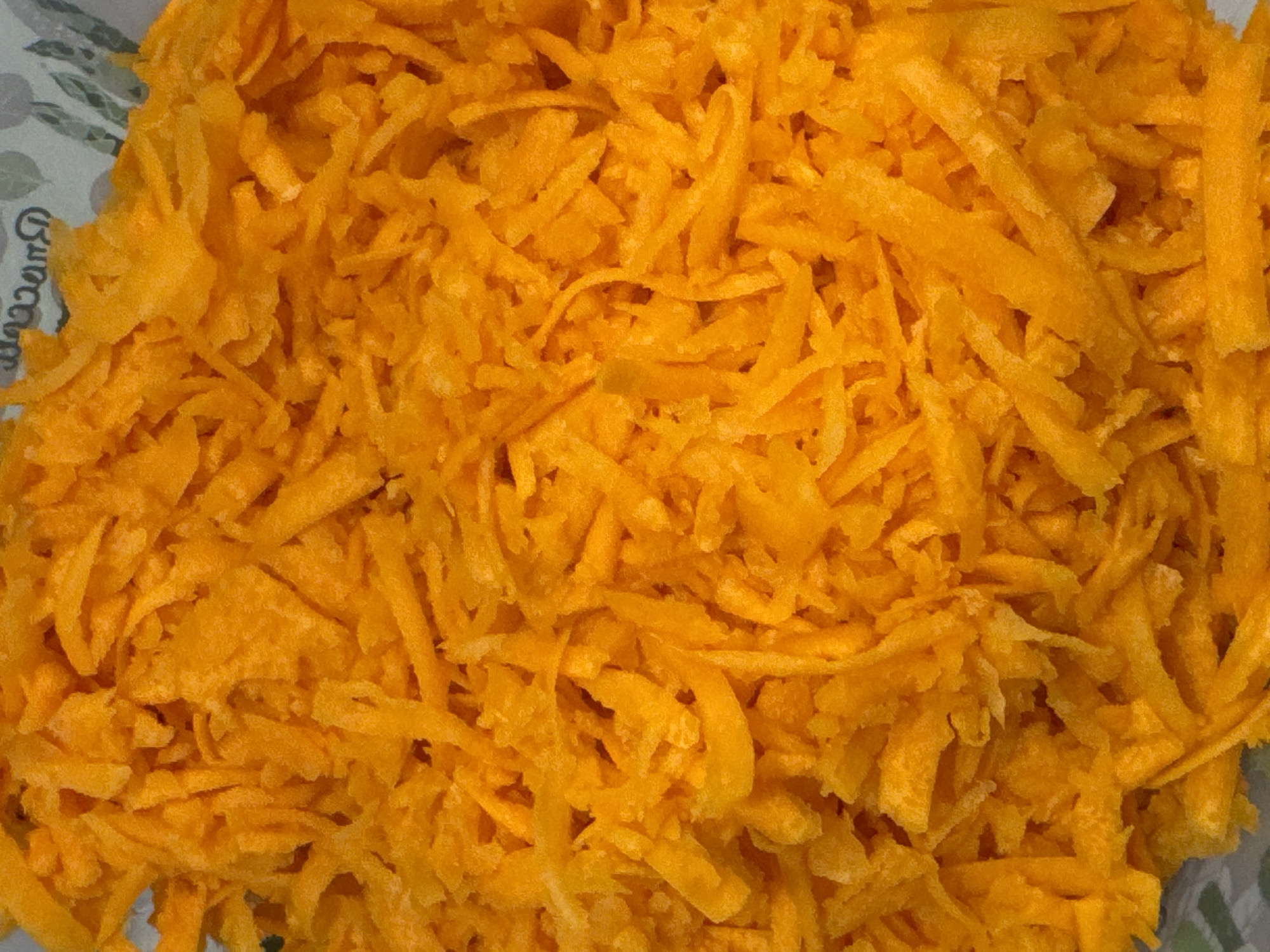 The image is a close-up, overhead shot of shredded carrots. The carrots are bright orange and appear to be finely grated, with thin strands of vegetable filling the frame. The texture is rough and fibrous. In the upper right corner of the image, a small portion of a patterned surface is visible, which appears to be a paper or fabric material with a faint green and gray design. The scene is domestic or culinary in nature and does not depict any specific urban environment, including Saratov, Russia.