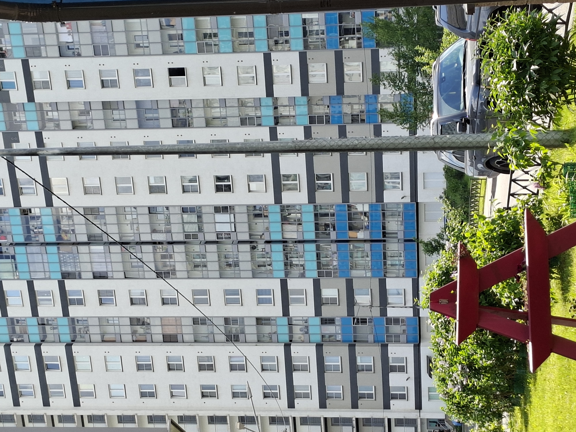 The image, taken in Saint Petersburg, Russia, displays a modern, multi-story residential building under bright daylight. The facade of the tall apartment block features a distinctive repeating pattern of light gray and dark gray panels, with numerous rectangular windows. Alternating bands of the building incorporate reflective or blue-tinted glass panels, adding a contemporary aesthetic. Some windows are open, suggesting residential use.
In the mid-ground, a dark-colored car is parked on what appears to be a paved street or parking area alongside a lush green strip of bushes and grass. A vertical metal pole stands prominently in the middle of the frame. The foreground is dominated by vibrant green foliage and a striking red, three-dimensional star-shaped sculpture, possibly a decorative element or a monument. No human activity is visible, and the scene appears static. There is no discernible text in the image.