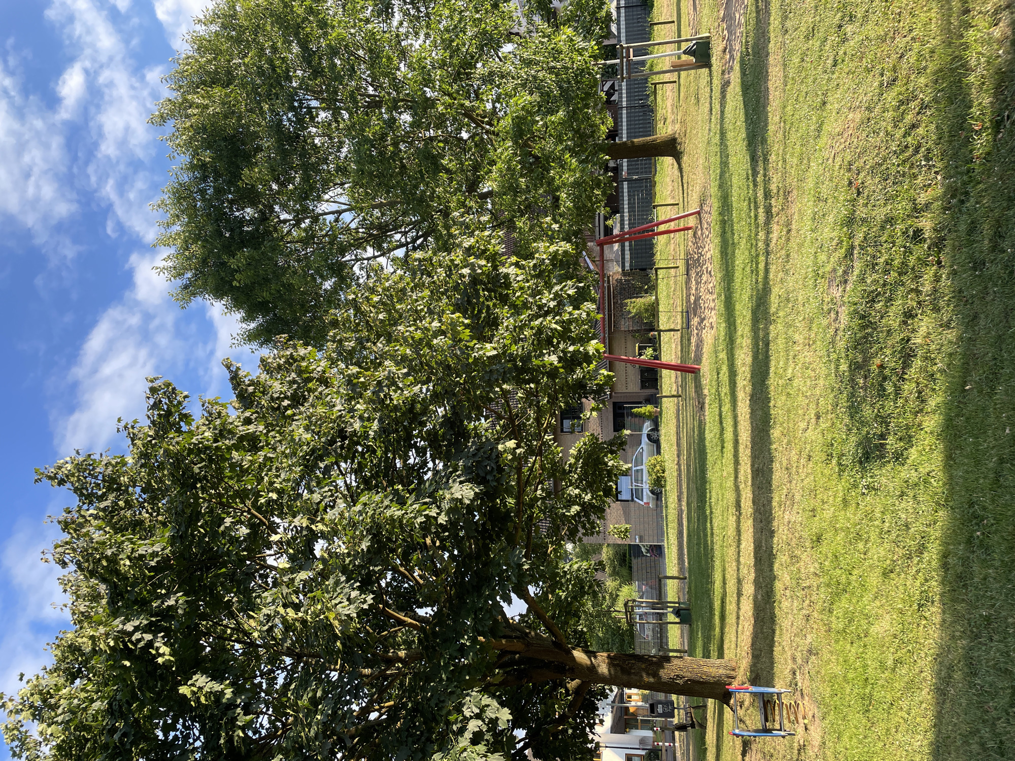 A sunlit playground is depicted, with two mature trees dominating the scene. The trees, with their full green foliage, cast shadows across the neatly mowed grass. In the background, a residential building and a parked silver car are visible, along with a fence. A red swing set with sandy ground underneath is situated between the trees. To the left of the swing set, a small blue and red play structure with two steps is positioned on the grass. The sky above is a vibrant blue, dotted with scattered white clouds. The image captures an outdoor scene in a residential area of Hennef, Germany, likely during daytime.