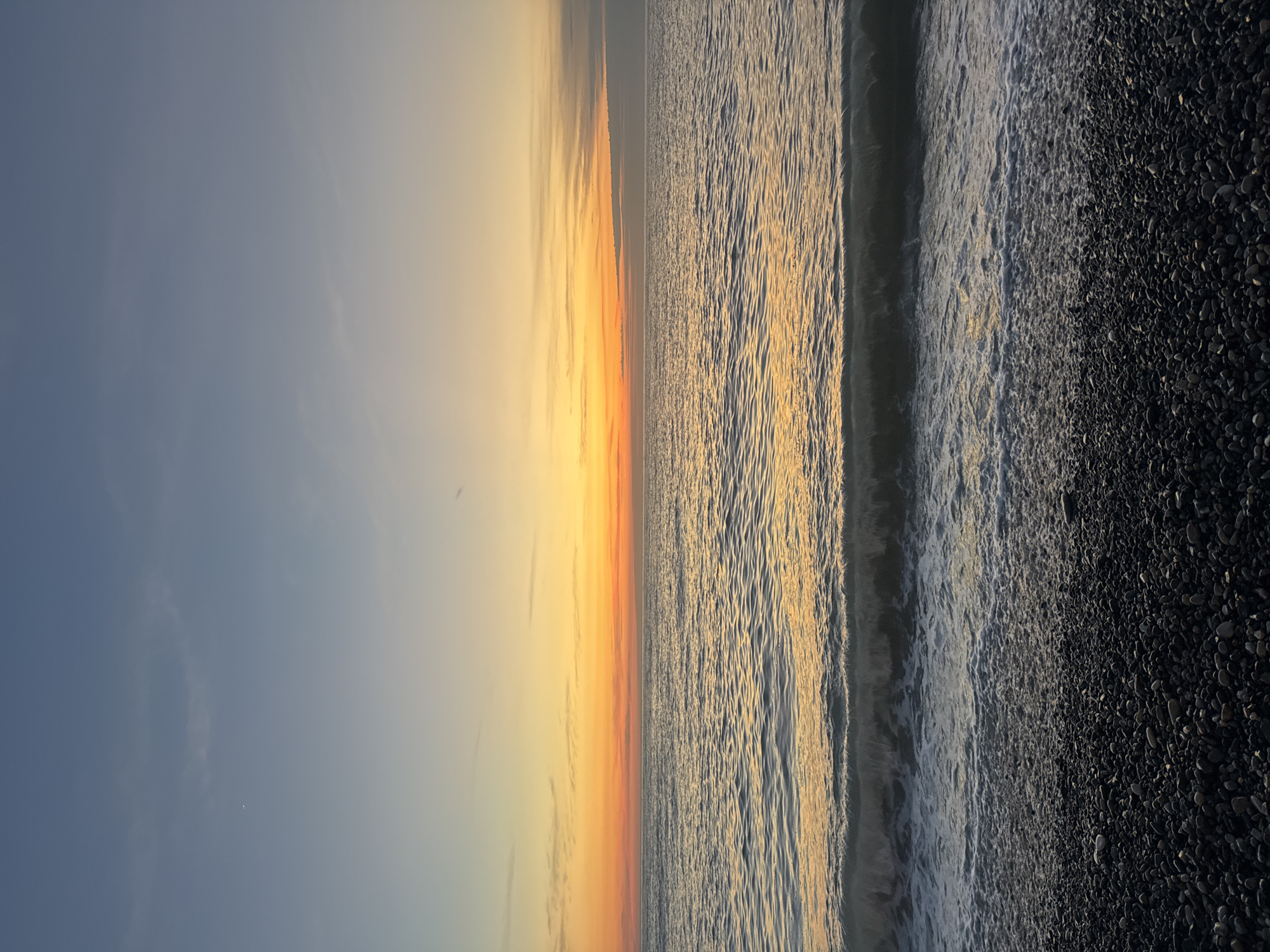 The image depicts a serene beach scene in Batumi, Georgia, captured at sunset. The left side of the frame showcases a gradient sky transitioning from a pale blue at the top to a warm orange and yellow hue near the horizon. The sea occupies the central portion of the image, its surface reflecting the colors of the sky. Gentle waves approach the shore, their foamy edges visible against the dark, pebbly beach on the right. The horizon line is clear and straight, dividing the sky and sea. No people are visible in the scene.