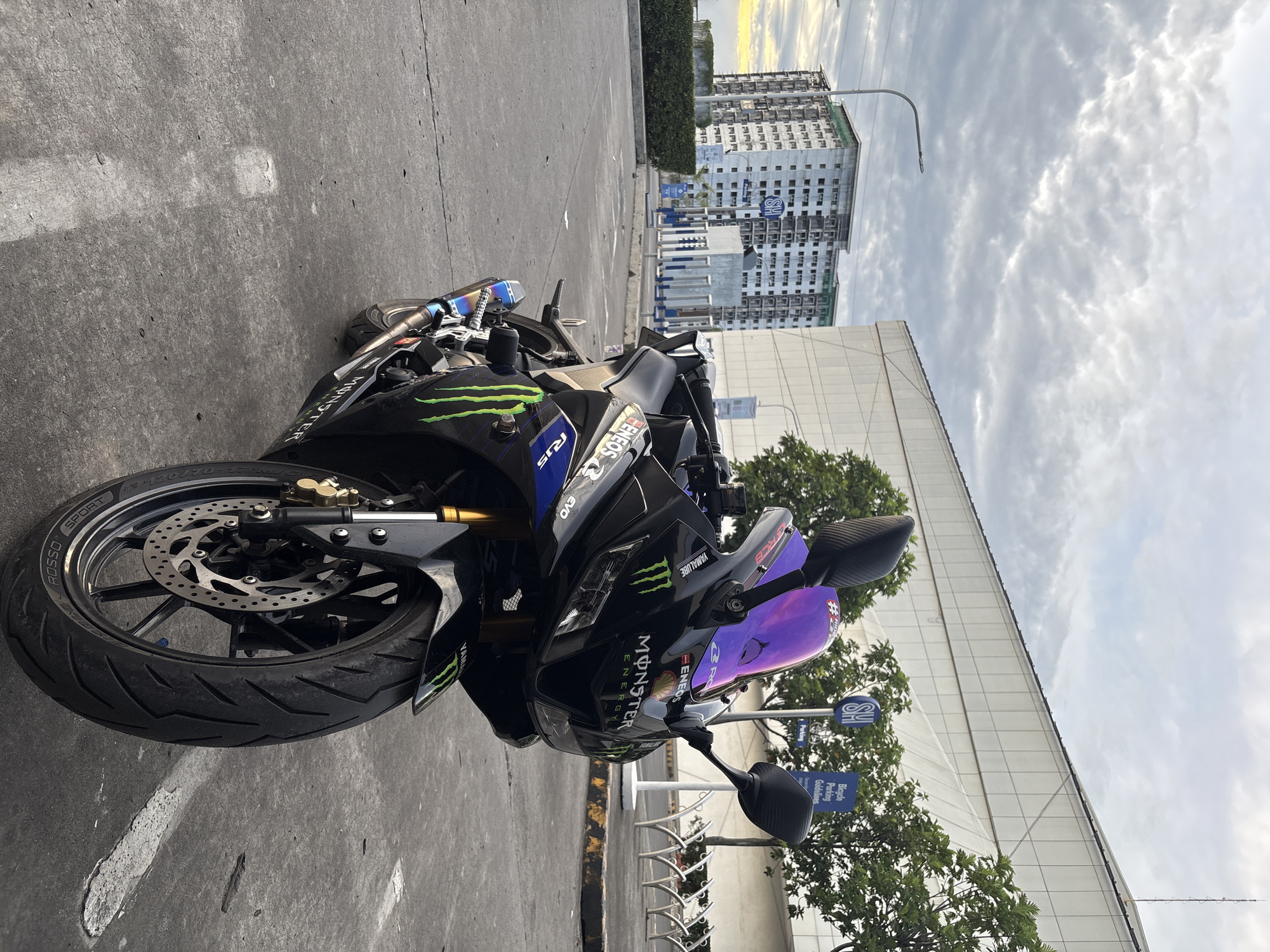 The image shows a black Yamaha motorcycle parked on a concrete surface. The bike is adorned with Monster Energy stickers and features a purple windshield. In the background, there's a building under construction with a "SM" logo visible, suggesting the location is likely near a SM shopping mall. Trees and a bicycle parking sign are also visible in the immediate background. The sky is overcast with grey clouds. Based on the SM logo, the location is likely in Davao City, Philippines, where SM malls are prevalent. The motorcycle appears to be parked in a designated parking area.