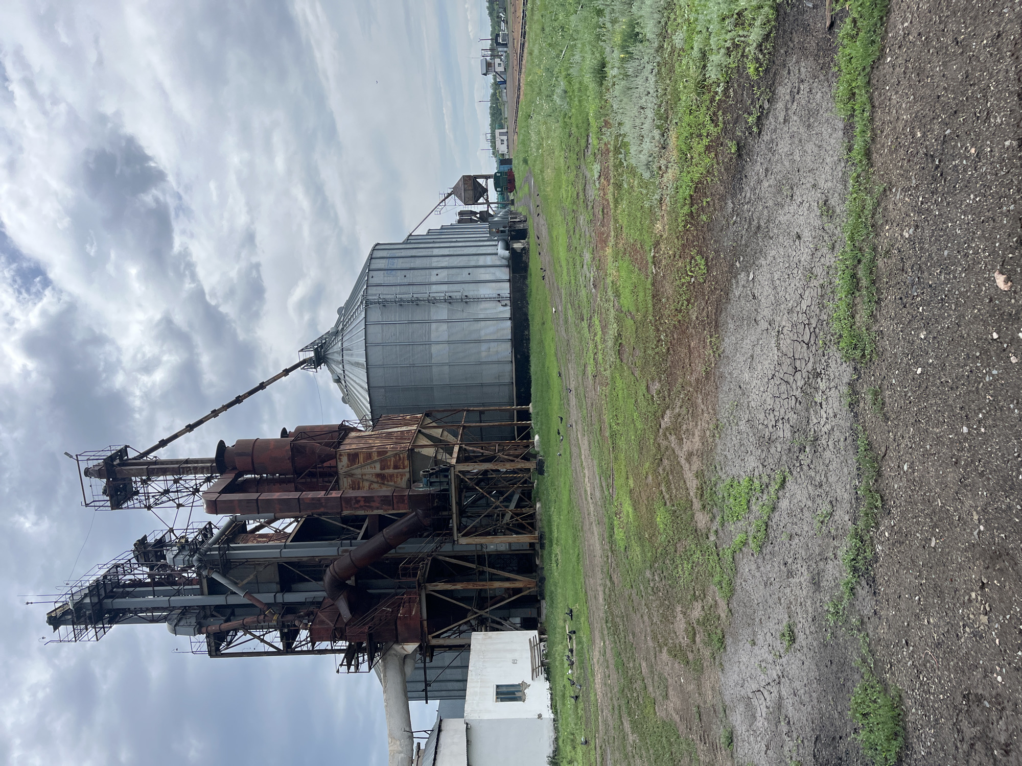The image depicts an industrial scene in the city of Progress, Kazakhstan. In the foreground, a dirt and gravel area with patches of dry, cracked mud and sparse green grass is visible. A small, white building with a single window and a wooden bench sits to the left. Adjacent to the building is a large, weathered industrial structure with multiple towers, rusty pipes, and metal platforms, likely part of a grain processing or storage facility. To the right of this structure is a large, silver, corrugated grain silo. Pigeons are scattered across the grassy area in front of the industrial complex. In the background, more industrial buildings, a railway line, and distant structures are visible under a cloudy sky.