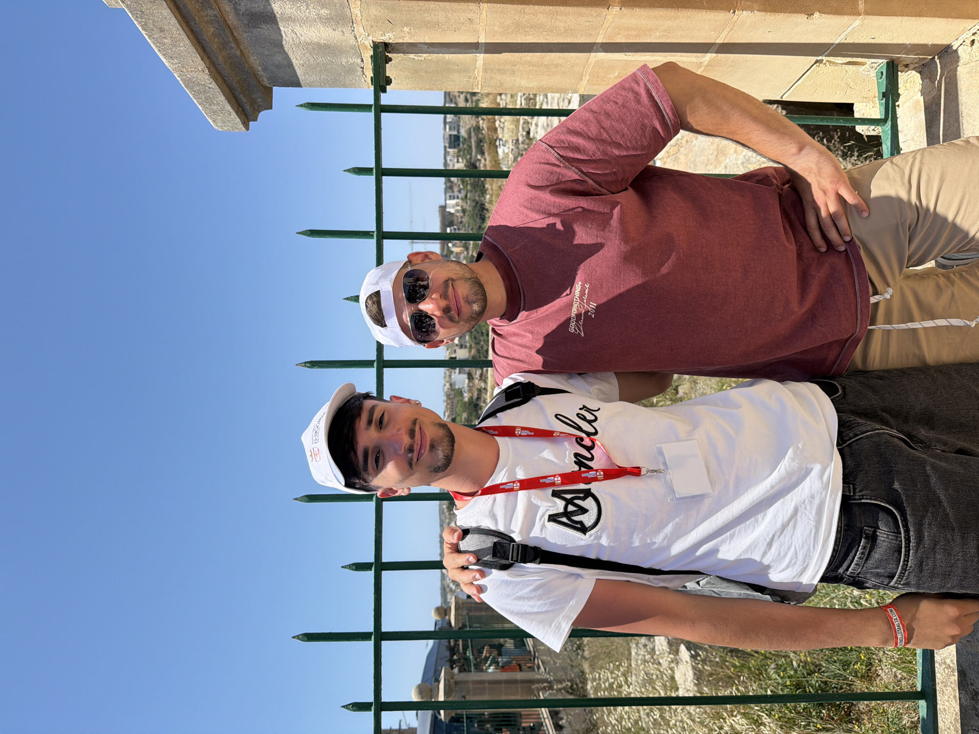 Two young men pose behind a dark green metal fence with spear-like tips. The man closer to the camera wears a white t-shirt with a black logo and lettering, a white baseball cap, black jeans, a backpack, and a lanyard with a tag. The man further back wears a burgundy t-shirt, white sunglasses, a white baseball cap, and tan pants. The sky is clear and blue. In the background, buildings are visible. The location appears to be in a city, although the specific city is unknown.