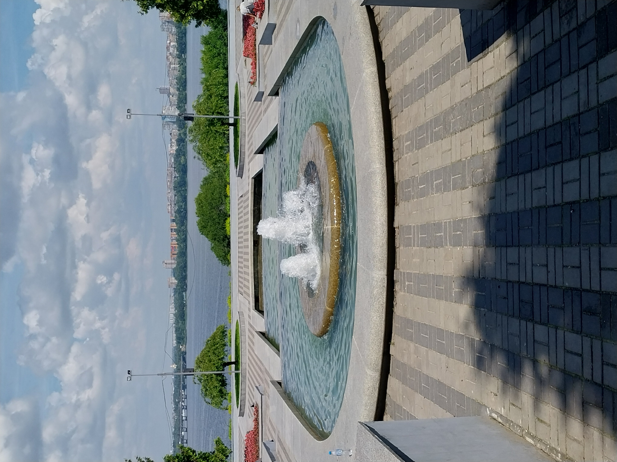 The image shows a view of a fountain in the city of Dnipro, Ukraine. The fountain is centrally located in a wide, shallow pool with a stone rim. The water in the pool is clear and rippling, with two jets of water shooting upwards from a circular stone structure in the center. Surrounding the pool is a paved walkway with a geometric pattern of gray and beige rectangular pavers. In the background, a wide river is visible, with a city skyline on the opposite bank. Trees and greenery line the riverbank. The sky above is partly cloudy, with patches of blue sky visible between white clouds. A single plastic bottle of water stands on the edge of the fountain's stone rim.