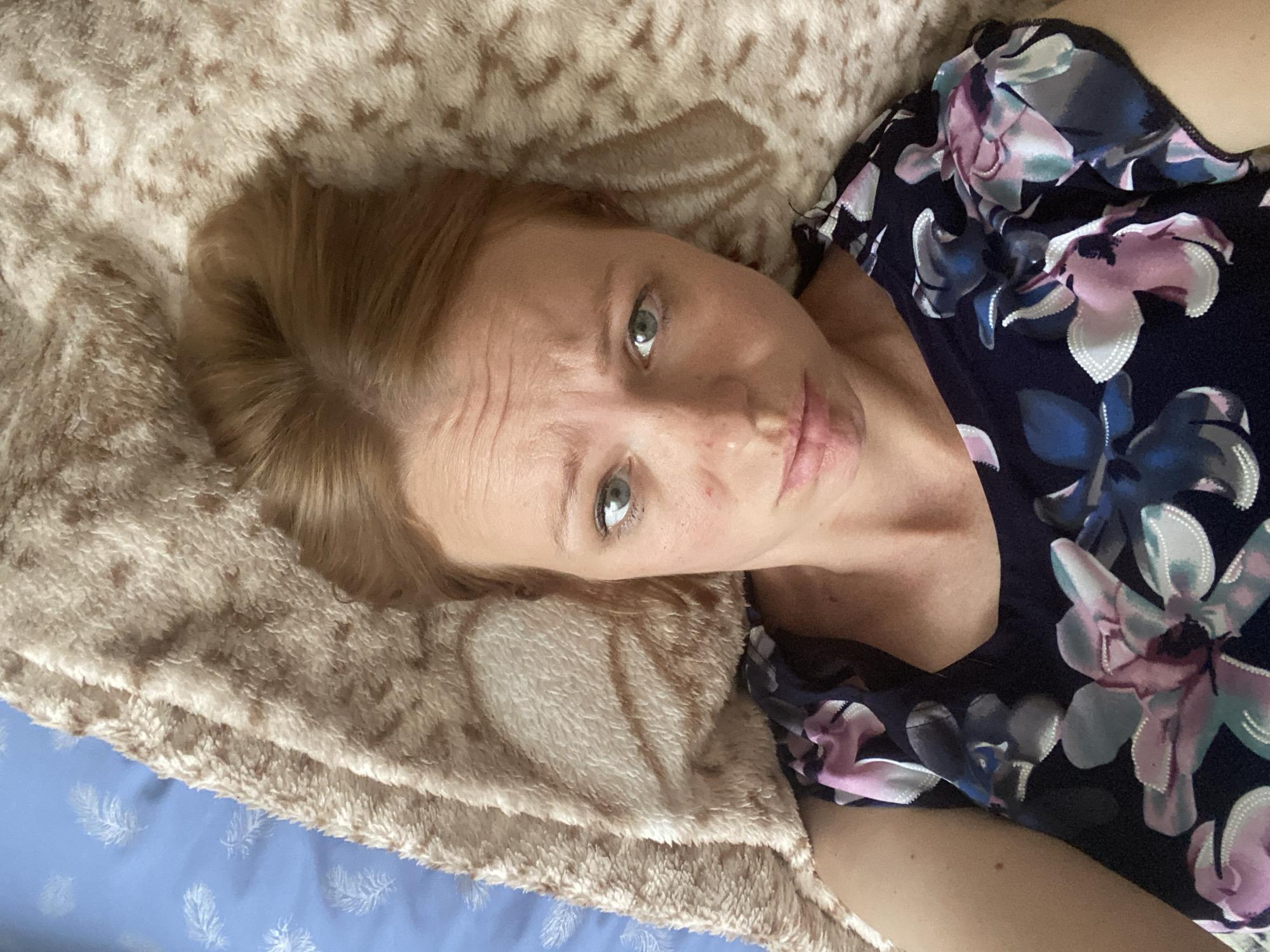 A fair-skinned woman with auburn hair is lying on a bed, looking directly at the camera. She is wearing a dark blue dress with a floral pattern of pink, white, and blue flowers. Her hair is pulled back loosely. The bed is covered with a fuzzy, beige blanket and a blue sheet with a white feather pattern. The lighting suggests an indoor setting, possibly a bedroom. There are no specific landmarks visible in the image that would indicate a location within Maykop, Russia.