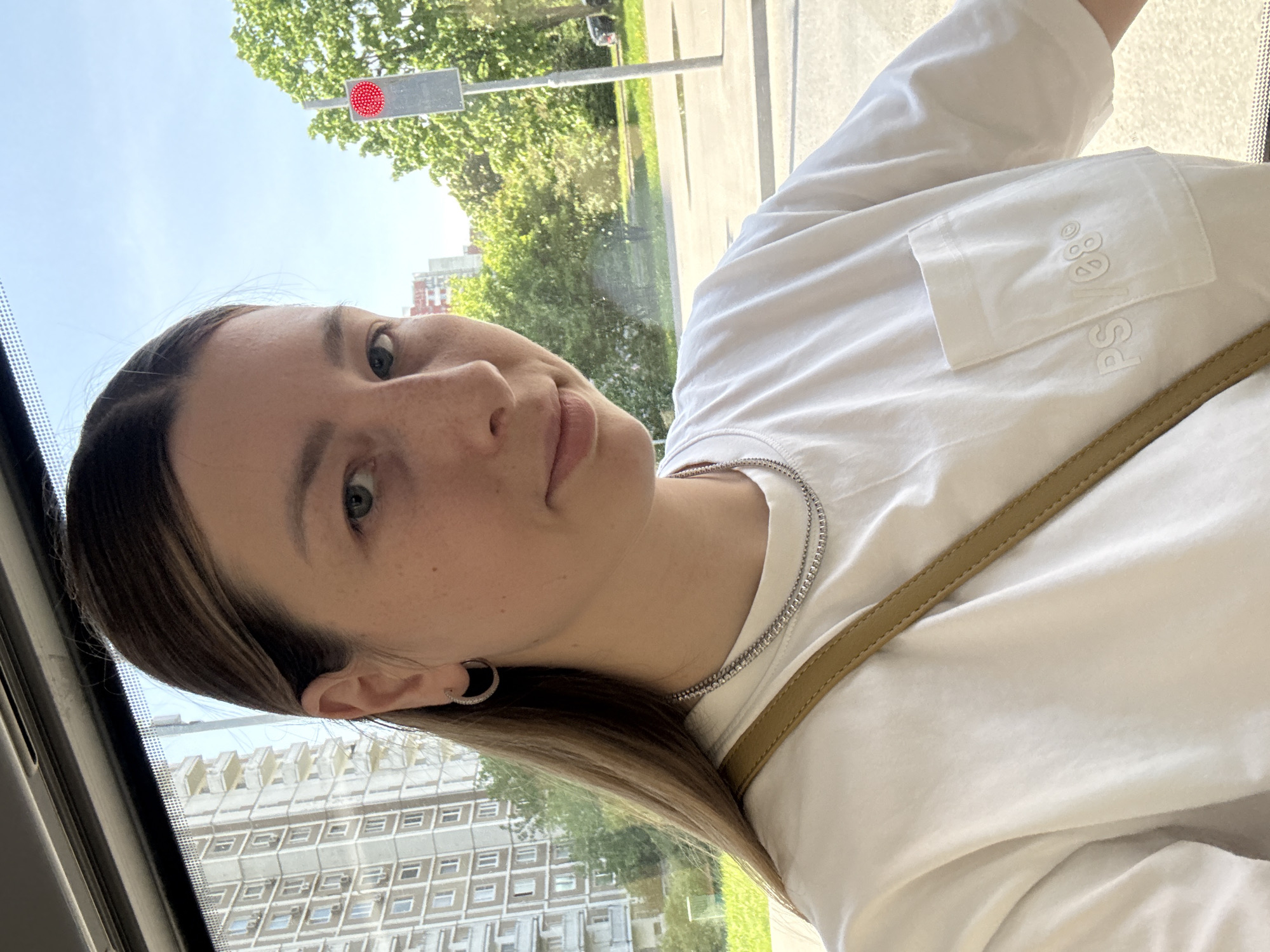 A young woman with long brown and blonde hair, wearing a white t-shirt and a silver chain necklace, is taking a selfie in what appears to be a vehicle. Through the window behind her, one can see a residential building with a repeating pattern of square windows and trees. There is a traffic light with a red light displayed. Based on the architectural style of the residential building in the background, this image was likely taken in Moscow, Russia.