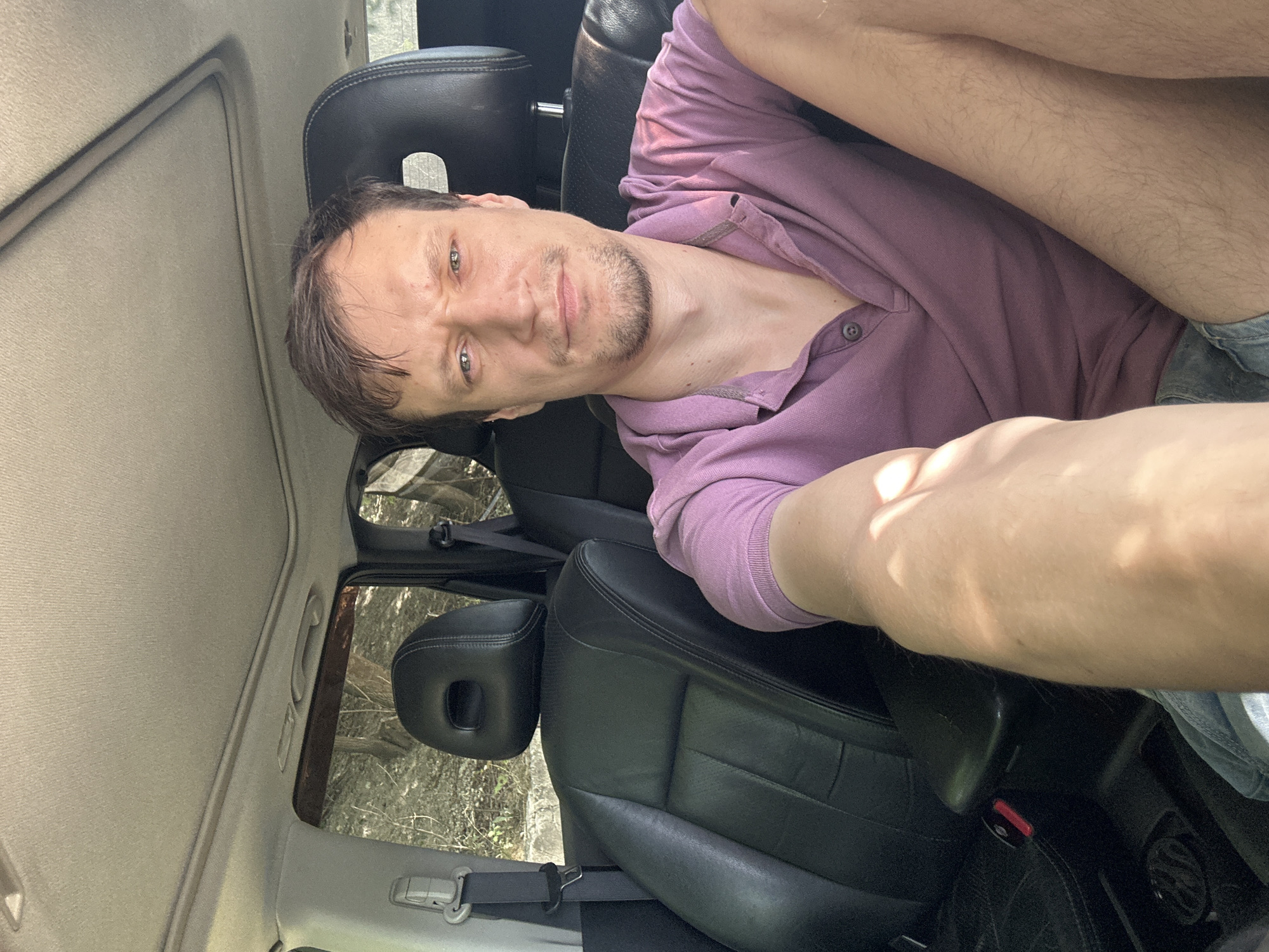 In Makiv, Ukraine, a man with short brown hair and stubble sits in the passenger seat of a car, looking directly at the camera with a slightly furrowed brow. He is wearing a purple polo shirt and denim shorts. The interior of the car is visible, featuring black leather seats, a sunroof, and a grey headliner. Outside the car windows, trees and foliage suggest a natural, possibly rural, setting. The lighting indicates it is daytime, with sunlight casting shadows on the man's arm and leg.