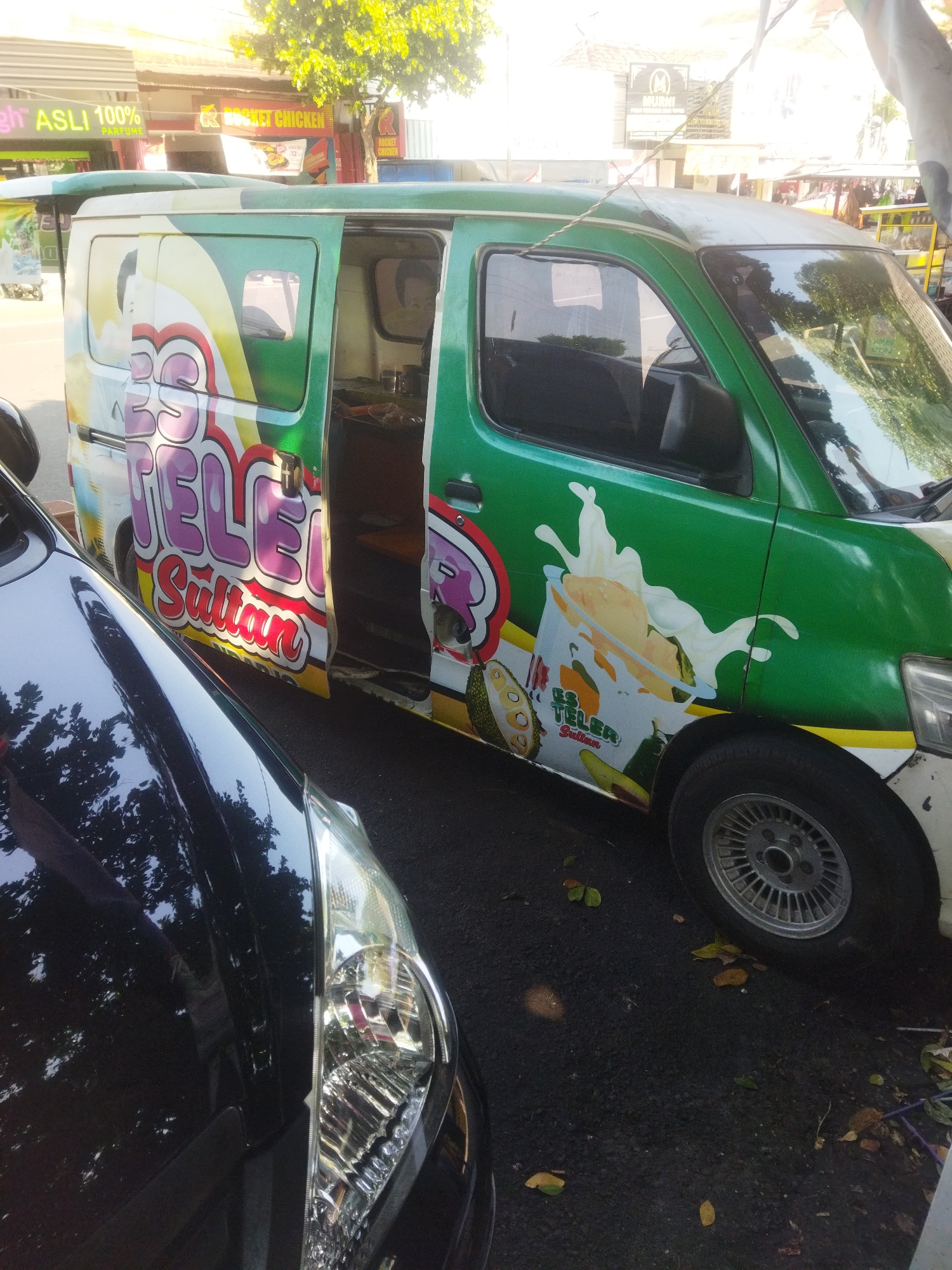 A green van with "Es Teler Sultan" branding is parked on an asphalt road. The van features colorful graphics of ice cream, fruit, and a person's silhouette. Its side door is open, revealing a counter and what appears to be food preparation supplies inside. A dark-colored car is parked to the left of the van, with its headlight visible. In the background, buildings with signs like "Rocket Chicken" and "MURNI" are visible, suggesting an urban commercial area. A leafy green tree is present above the buildings, and the scene is illuminated by daylight, casting shadows on the ground. The overall impression is a street scene in a commercial district in Wonogiri, Indonesia, with a food vendor vehicle as the primary subject.