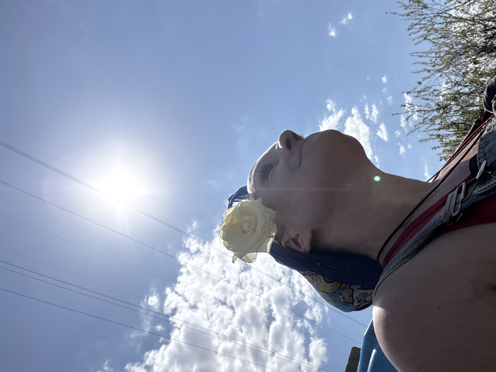 The image shows a person looking up toward the sun in a cloudy sky. A white rose is tucked behind their ear. Electrical power lines are visible in the sky. The location is identified as Chaikovsky, Russia.