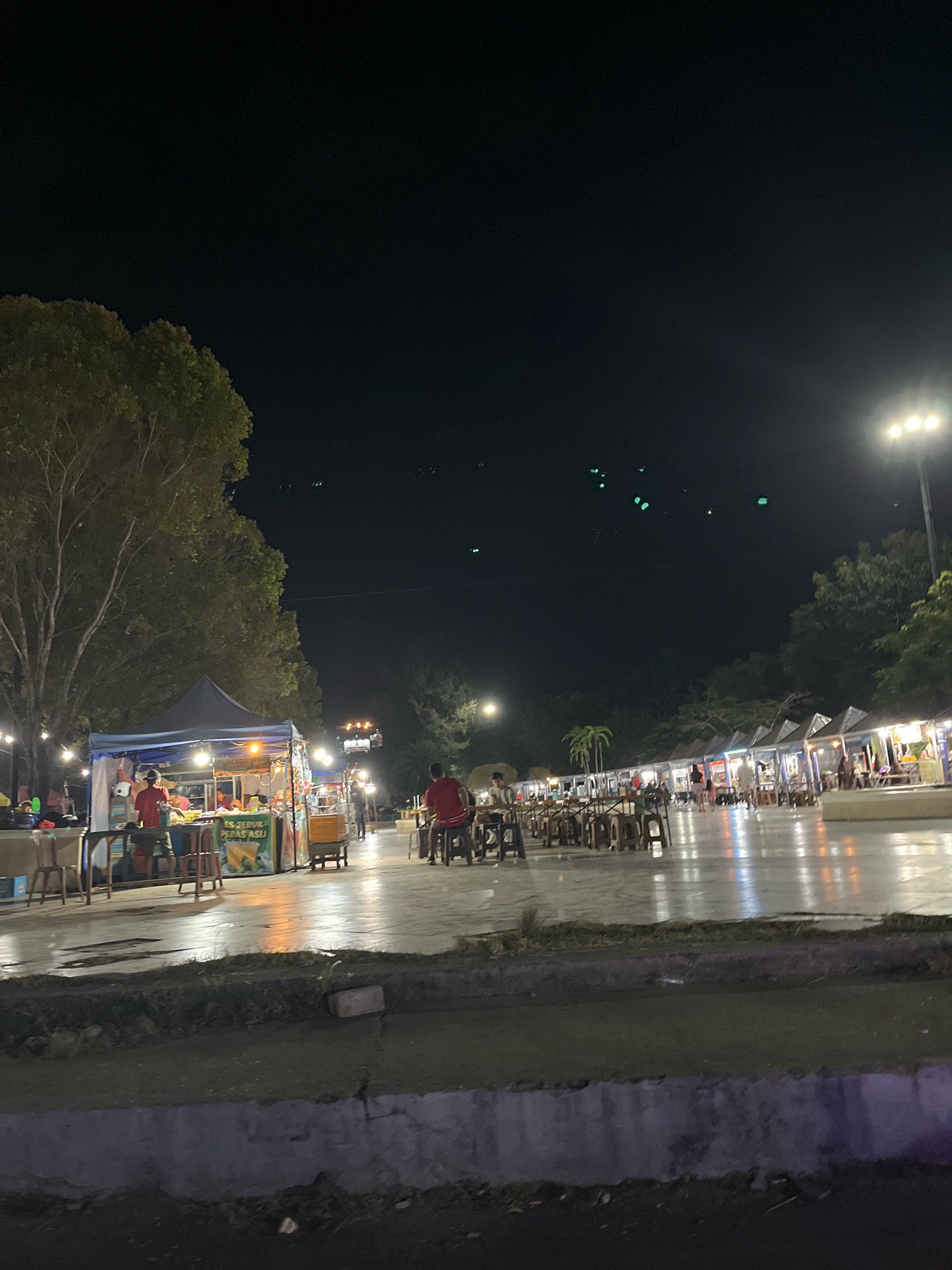 This is a nighttime street scene at a food market in Kendari, Indonesia. The image captures a lively atmosphere with numerous food stalls set up along a paved area. Several individuals are visible, some tending to the stalls, while others are seated at tables, presumably eating or socializing. The stalls are illuminated by overhead lights, creating a warm glow that reflects off the wet pavement. In the background, trees and palm trees line the perimeter of the market, with streetlights and a tall pole light illuminating the area. The sky is dark, indicating it is well after sunset. The overall impression is one of a vibrant night market, bustling with activity and a variety of food vendors.