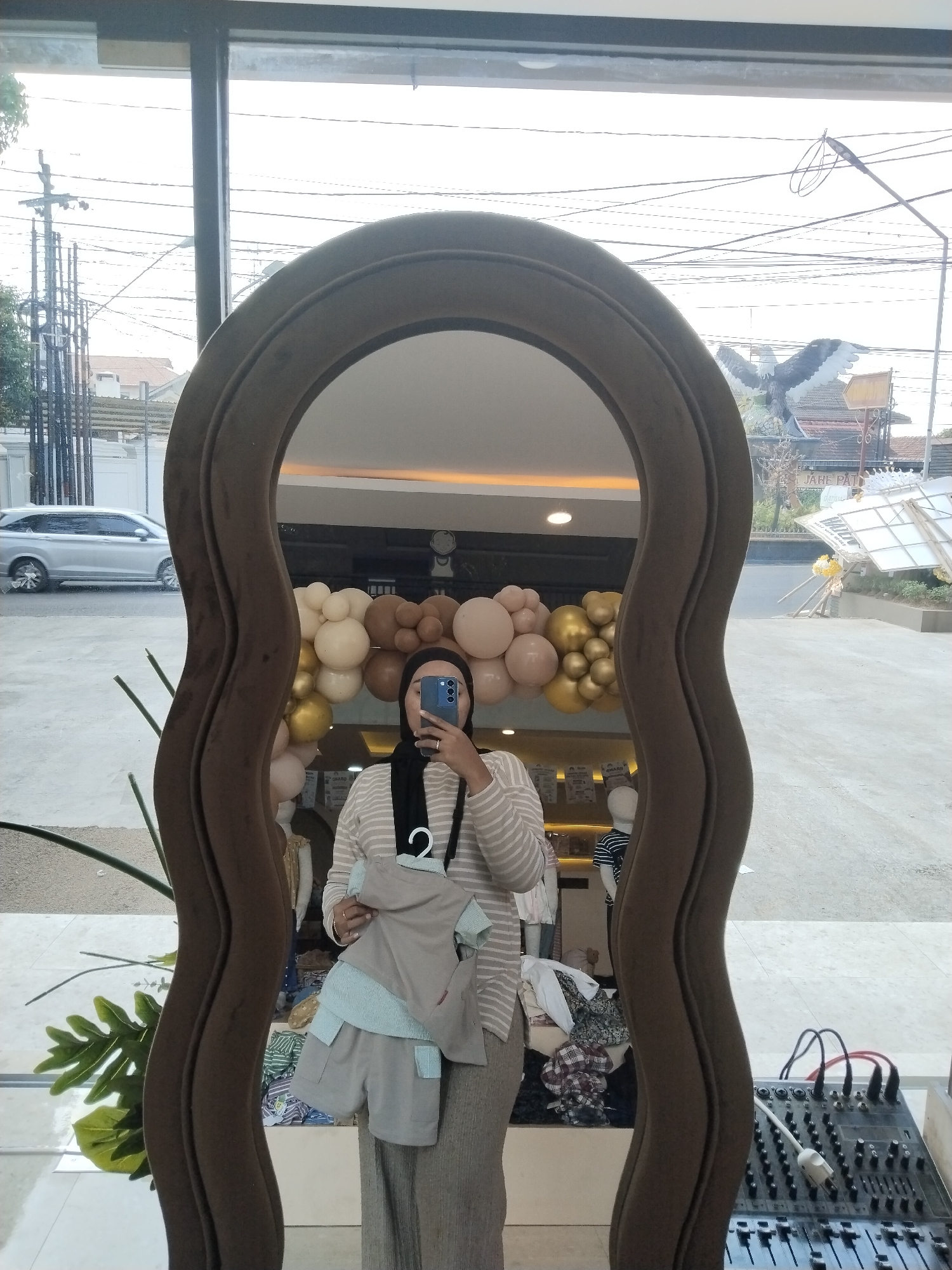 A person wearing a black hijab and a striped sweater stands in front of a large, ornate mirror, taking a selfie. They are holding a grey and light blue baby outfit. The mirror reflects a shop interior with balloons, mannequins, and clothing displays. Outside the shop, visible through a large window, is a street scene with a silver car, power lines, and buildings. The image appears to be taken in front of a shop on Jalan Raya Pati-Juana in Kecamatan Pati, Indonesia, likely near the Plaza Pragolo. A sound mixer is visible in the lower right corner of the frame.
