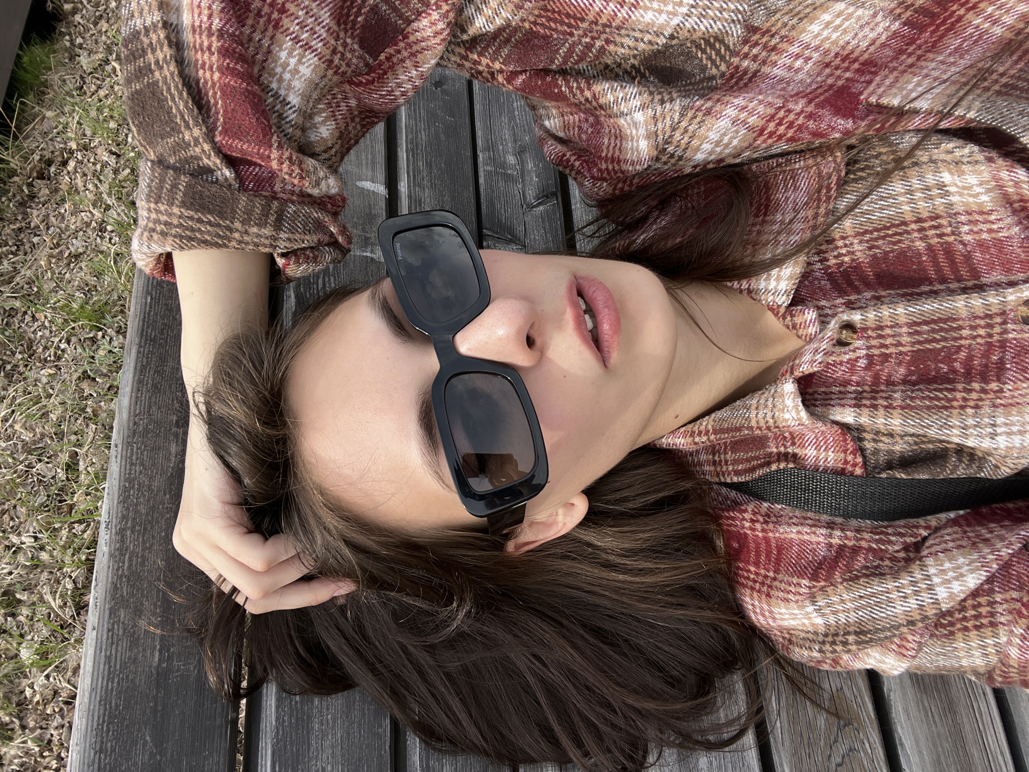 A young woman is lying on a wooden bench outdoors, wearing sunglasses and a plaid shirt. Her head is tilted towards the camera, with her right hand touching her hair. The bench appears to be in a park or similar outdoor setting, with grass and other vegetation visible in the background. The image was taken in the city of Krasnoyarsk, Russia.