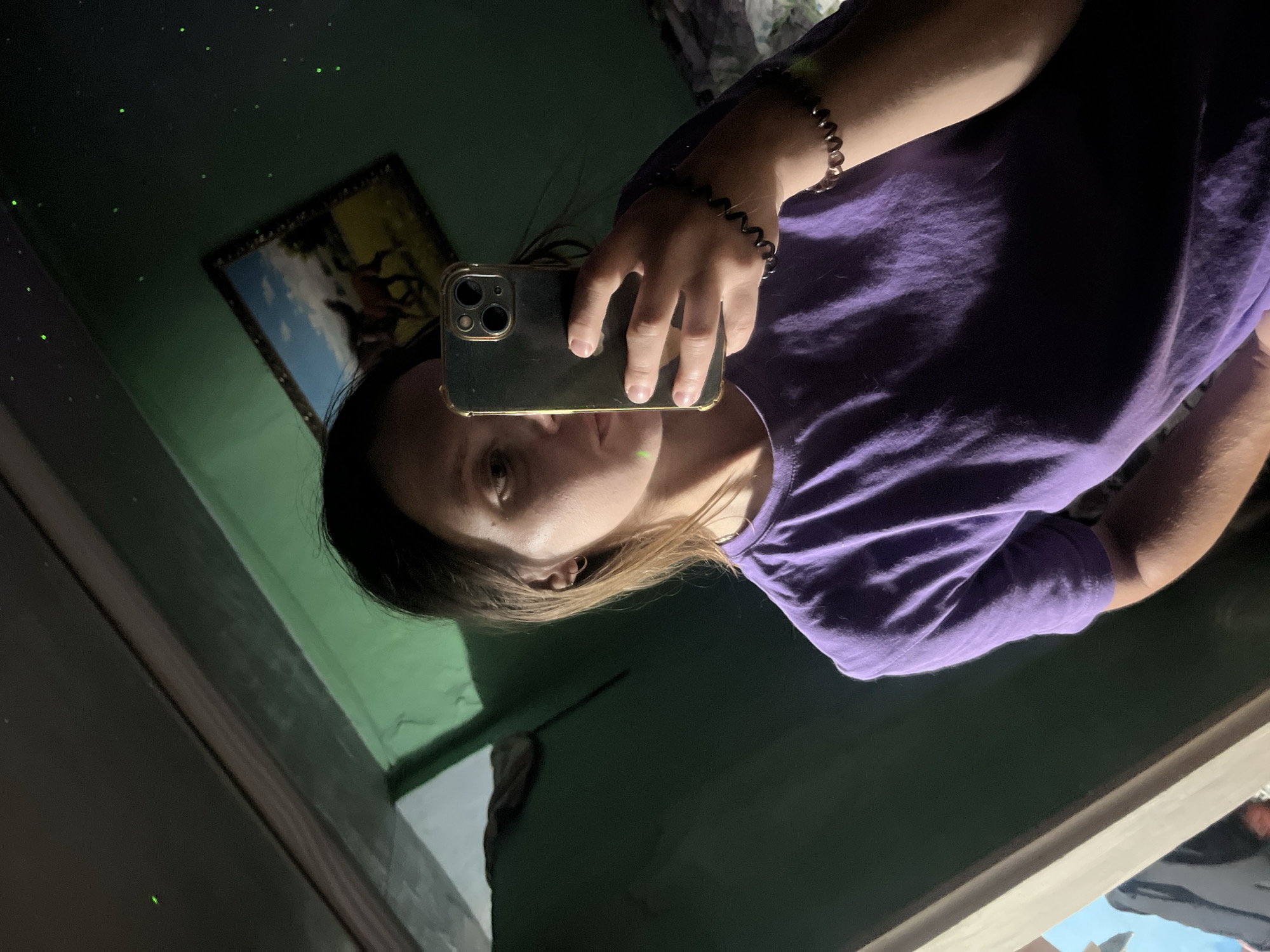 A woman is taking a mirror selfie in a room. She is wearing a purple t-shirt and has a bracelet on her left wrist. In the mirror, a framed picture of horses is visible on the wall, along with green light effects. The room appears to be dimly lit.