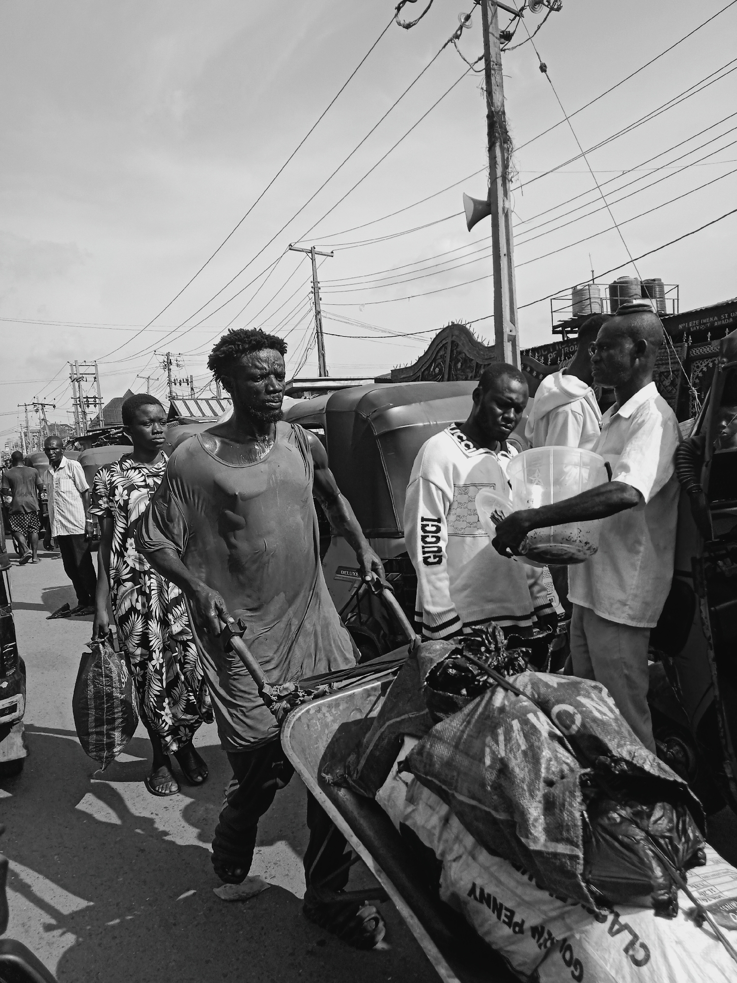 This black and white photograph depicts a bustling street scene in Onitsha, Nigeria, under bright daylight conditions. The central focus is a visibly sweaty or wet man with curly hair and a beard, pushing a heavy wheelbarrow filled with sacks. He wears a loose, dark garment over his bare chest, indicating manual labor.
Behind him, a woman in a patterned dress walks purposefully, carrying a bag. To the right, several other men are present, including one wearing a white shirt who holds a transparent plastic bucket, seemingly interacting with another man in a "GUCCI" branded sweater.
The environment is characterized by a dense network of overhead power lines strung between wooden poles, one of which features a loudspeaker. Partially visible auto-rickshaws (tricycles) and distant buildings with water tanks on their rooftops suggest a busy urban or market setting.
Visible text includes "GUCCI" on a man's sweater, "DELUXE" on a vehicle, and "GOLD PENNY" and "CLAS" on the sacks in the wheelbarrow. A sign on a building in the background reads "N0 1 EZE IWEKA ST OZ LAYOUT AWADA," providing a specific location context within Onitsha. The image captures a moment of everyday activity and commerce in a vibrant street.