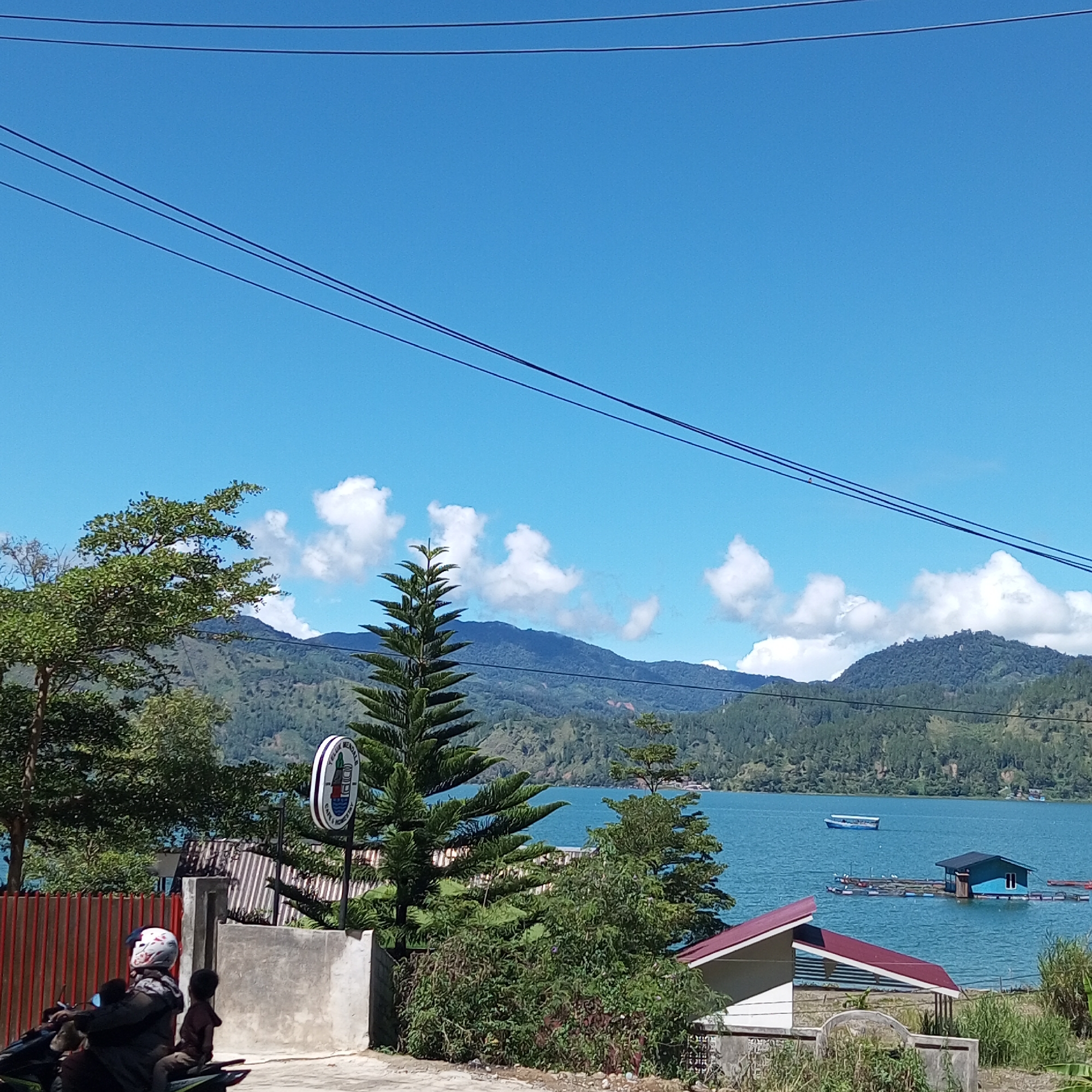 In the city of Kalalengkio, Indonesia, a vibrant scene unfolds under a clear blue sky with scattered white clouds. The foreground is dominated by a paved road where a person on a motorcycle, wearing a white helmet with a red and black design, is accompanied by a child. To the left, a red metal fence runs horizontally.
Further into the scene, a tall evergreen tree stands prominently, its branches reaching towards the sky. Behind it, a large body of water, likely a lake, shimmers with a bright turquoise hue. In the distance, rolling hills covered in dense green forests form a majestic backdrop, with layers of mountains receding into the haze.
On the water, several structures are visible. A small blue boat navigates the calm surface. Closer to shore, a blue-roofed floating structure with several compartments is anchored. Another, smaller floating structure with multiple colored buoys is also present.
To the right, a white building with a red roof is partially visible, suggesting a dwelling or a small establishment. The overall atmosphere is one of natural beauty and tranquility, with a touch of human presence.