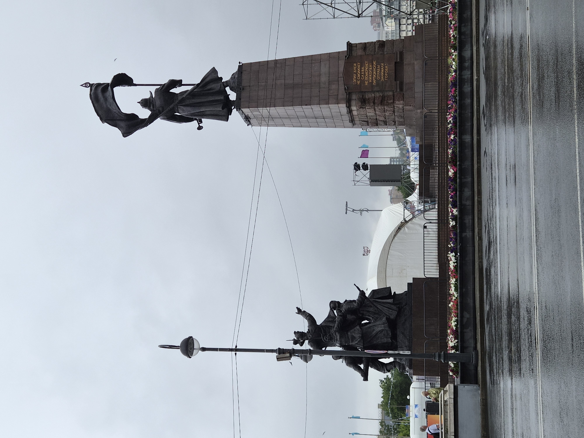 The image depicts the "Brave Hearts" monument located in Vladivostok, Russia. The monument features two bronze statues. The larger statue on the right depicts a figure holding a large flag. The smaller statue on the left shows a group of figures in dynamic poses. Both are set against a backdrop of a gray, overcast sky. In the foreground, a wet asphalt road reflects the scene, indicating recent rain. A lamppost stands between the two main statues. Behind the monument, there are tents, flags, speakers, and scaffolding, suggesting a public event or construction. A low flower bed runs along the base of the monument. Two individuals are visible in the lower left corner of the image, standing near a concrete barrier. The inscription on the base of the larger monument is visible, though the text is partially obscured. The overall atmosphere is somber due to the weather.