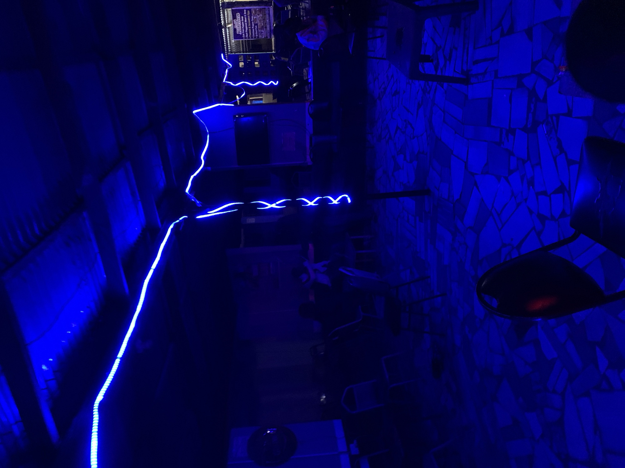 This image depicts an indoor or covered outdoor entertainment or dining venue, illuminated predominantly by blue LED strip lighting, located in New Stadium, Nigeria. The ceiling consists of corrugated material, from which blue LED strips are suspended. One strip forms a long, straight line across the upper left, while others on the right display a more undulating, decorative pattern. The flooring is a mosaic composed of irregularly shaped, light-colored tiles, appearing blue under the ambient light.
Multiple dark-framed chairs with lighter-colored seats are arranged in rows on the left side, some appearing unoccupied. In the mid-ground, towards the right, several individuals are seated at small, dark tables. One person on the far right is wearing a light-colored top. Further back on the right, a menu board is visible, labeled "Kitchen" and listing food items such as "Gala & Pepper Soup," "Assorted Portioned Meat & Soup," "Plantain & Toasted Eggs," "Ghana Jollof Rice," and "Spaghetti." A dark, circular object, possibly a speaker, is mounted on a wall or panel on the far left. A dark screen, resembling a television, is mounted on a background structure in the mid-right. A prominent dark chair is visible in the foreground, bottom-right.