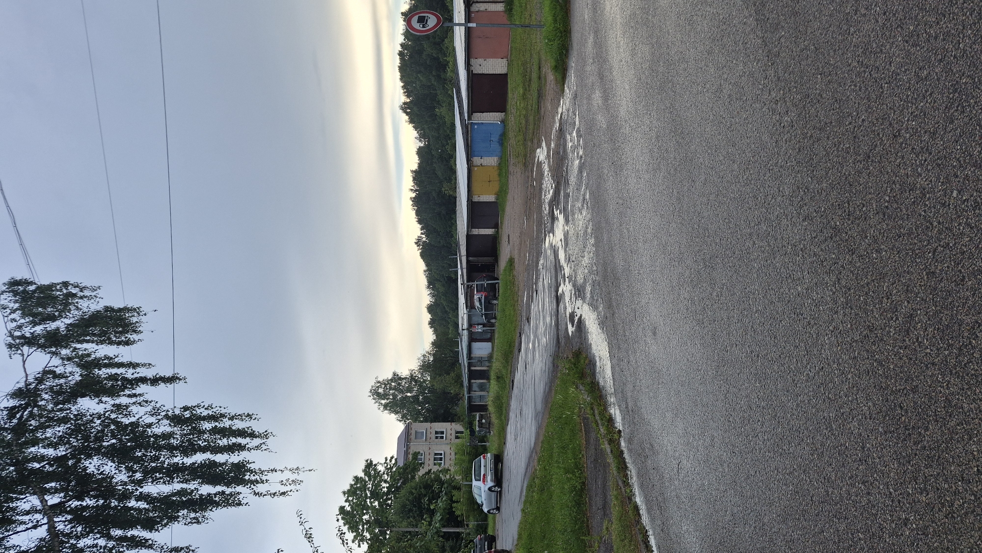 In Krauja, Latvia, a slightly overcast sky hangs above a residential area. A row of garages, some with colored doors (yellow, blue, red), stretches across the middle ground. A sign depicting a truck with a red circle and line through it, indicating no trucks, stands to the right of the garages. A silver car is parked near the garages, and another car is visible on the road. To the left, a multi-story building with multiple windows is surrounded by trees, and a large tree with hanging branches partially obstructs the view of the sky. The road in the foreground is wet and shows signs of puddles, suggesting recent rain.