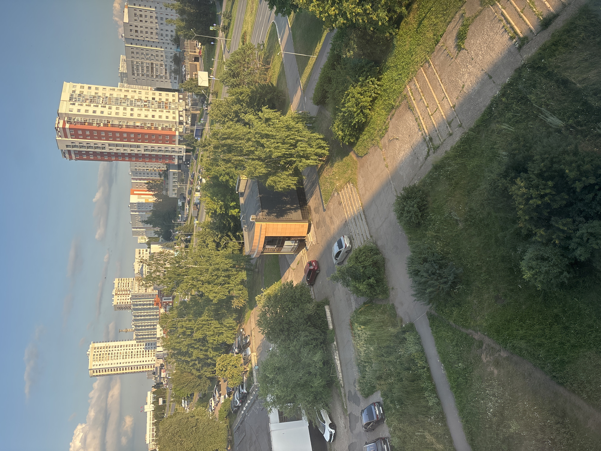 This media file captures an aerial view of a residential area in Naberezhnye Chelny, Russia. The scene is dominated by tall, multi-story apartment buildings, some with red and white facades, lining a street. The time of day appears to be late afternoon or early evening, suggested by the long shadows cast by the buildings and trees. The sky is clear with a few wispy clouds.
The urban landscape is interspersed with abundant greenery, including mature trees and grassy areas. Several cars are visible parked along the streets and in parking lots, indicating a typical day in a populated area. A small, modern building with a wooden facade is also present, situated between a road and a grassy embankment. A concrete pathway with raised sections runs alongside a green slope. The overall impression is one of a functional, well-established residential district bathed in the warm light of the setting sun. No visible text is present in the image.
