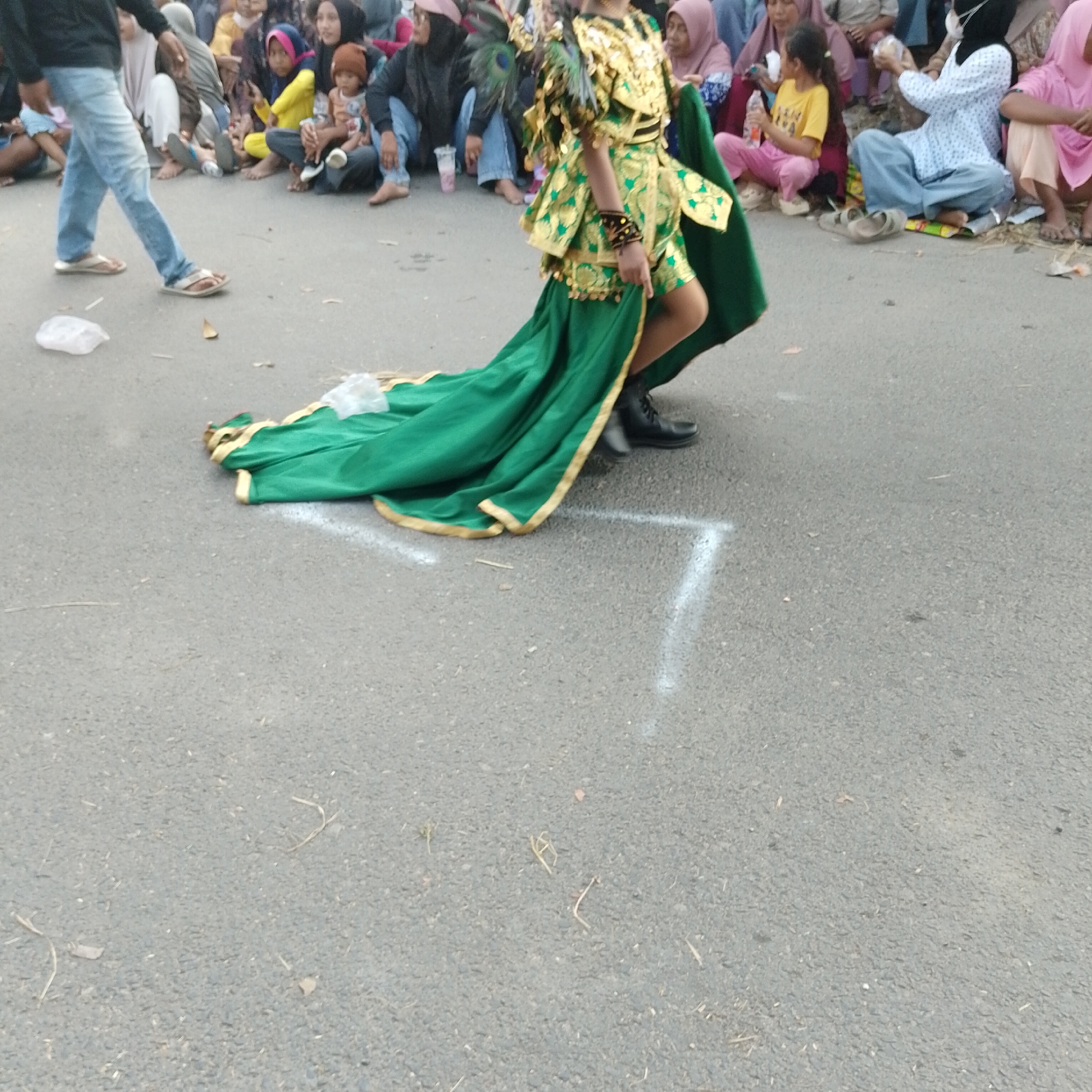 The image depicts a street scene in Palang, Indonesia, likely during a local event or festival. In the foreground, a person dressed in an elaborate, gold and green costume with a long, flowing train is walking on an asphalt road. The costume features intricate gold embellishments and feather accents. The person is wearing black boots. Behind this central figure, a crowd of spectators is seated on the roadside, observing the event. They are dressed in casual attire, with many wearing hijabs, indicating a predominantly Muslim population. Children are present among the spectators, some being held by adults. The environment appears to be an outdoor public space, with the road marked with white lines that might delineate a performance area or pathway. Debris, such as plastic bags and dry leaves, are scattered on the road and the sides, suggesting a lively, possibly informal gathering. The overall impression is one of a community celebration or parade taking place in a typical Indonesian street setting.